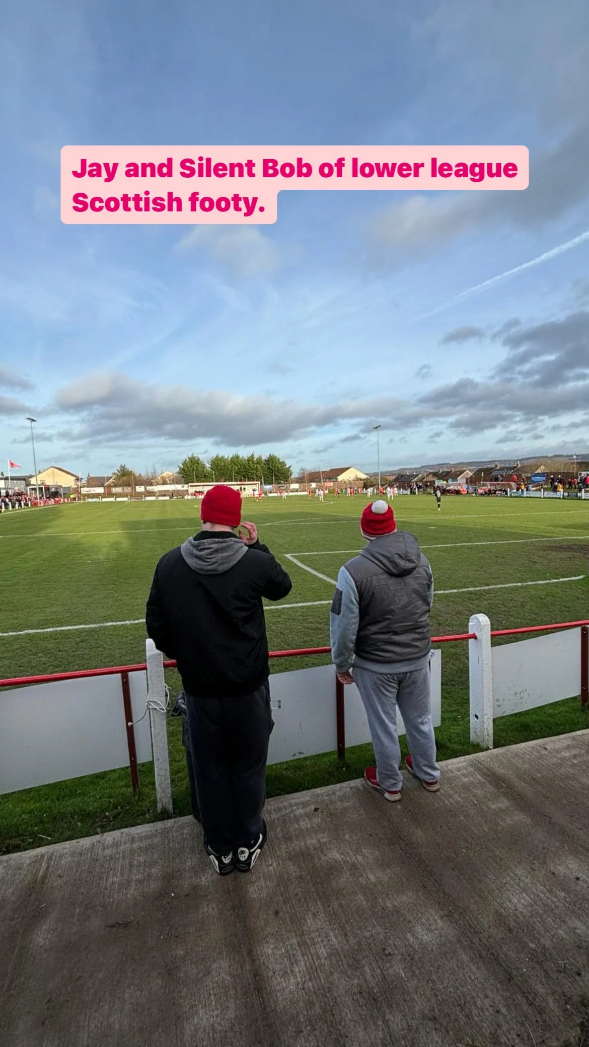 Two men wearing red Scottish football hats and casual clothing watching a soccer game on a green field under a cloudy sky.