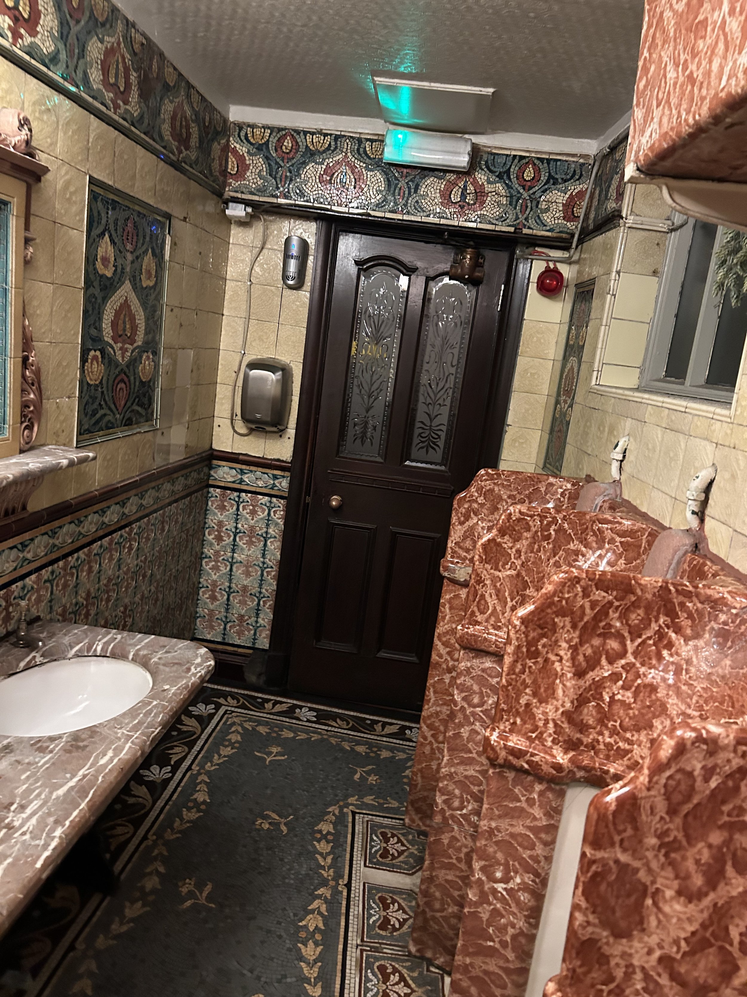 Public restroom with ornate tile walls, a marble countertop with a sink, several pink marble-patterned toilet stalls, a dark wooden door, vintage-style wallpaper, a small mirror, and a window with blinds.