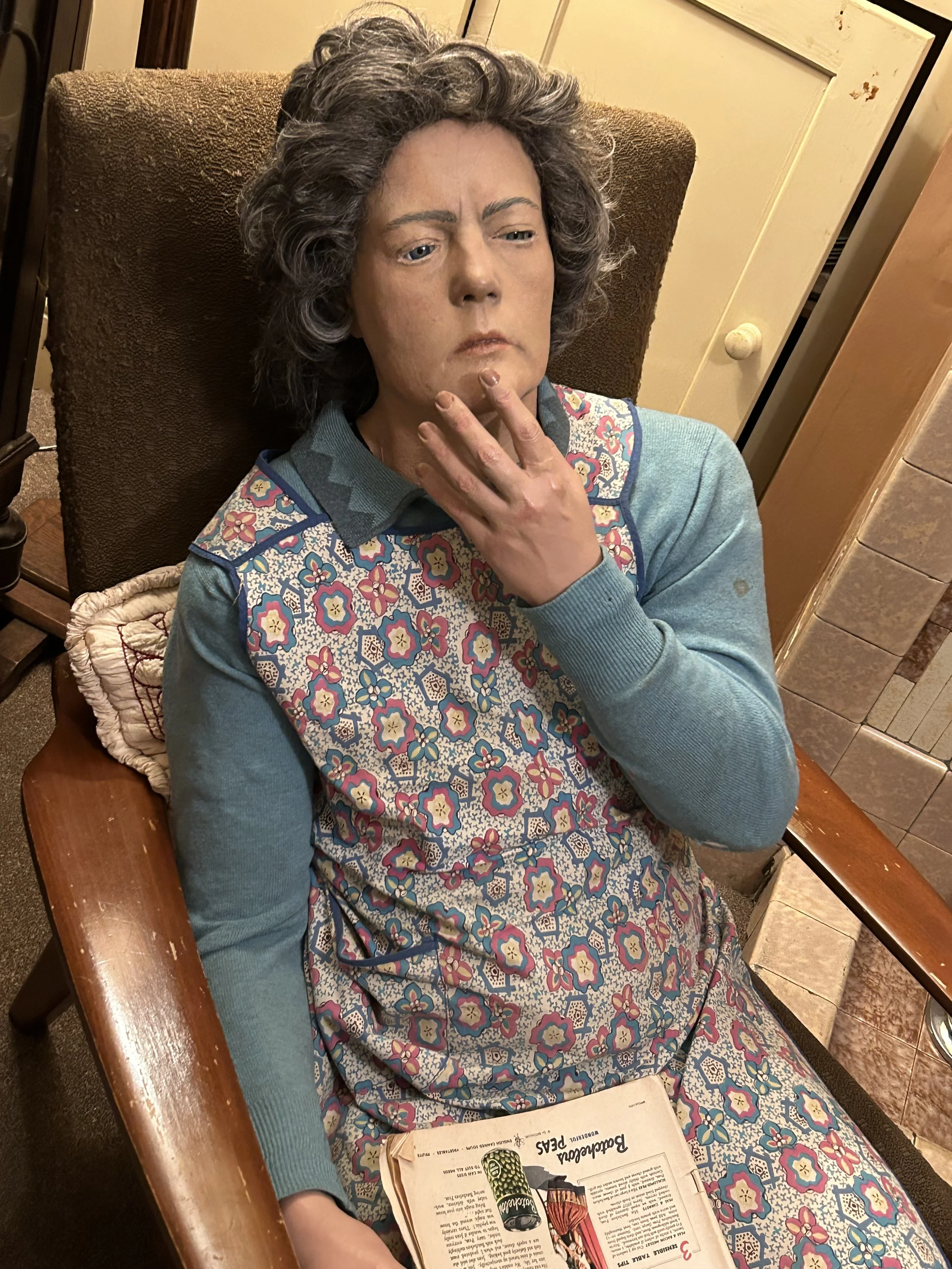 A woman with gray hair, dressed in a floral-patterned dress over a teal long-sleeve shirt, is sitting in a brown armchair. She is touching her chin with her right hand and has a thoughtful expression. An open magazine and a green glass bottle are on 