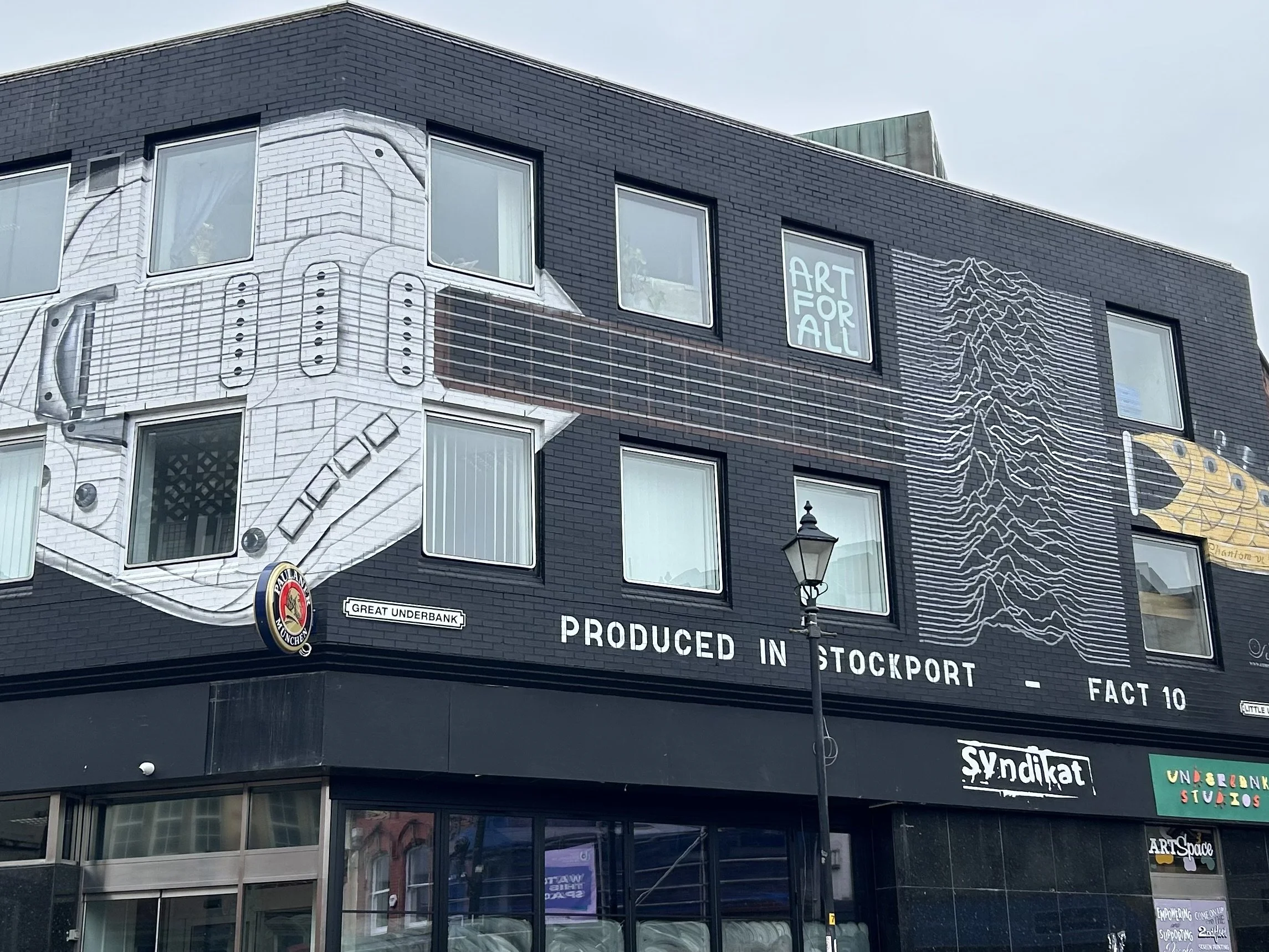 Murals and signs on a building on a city street, including artwork with geometric patterns and text, a street sign reading 'Great Underbank', and signs for 'Syndikat' and 'Art Space'.