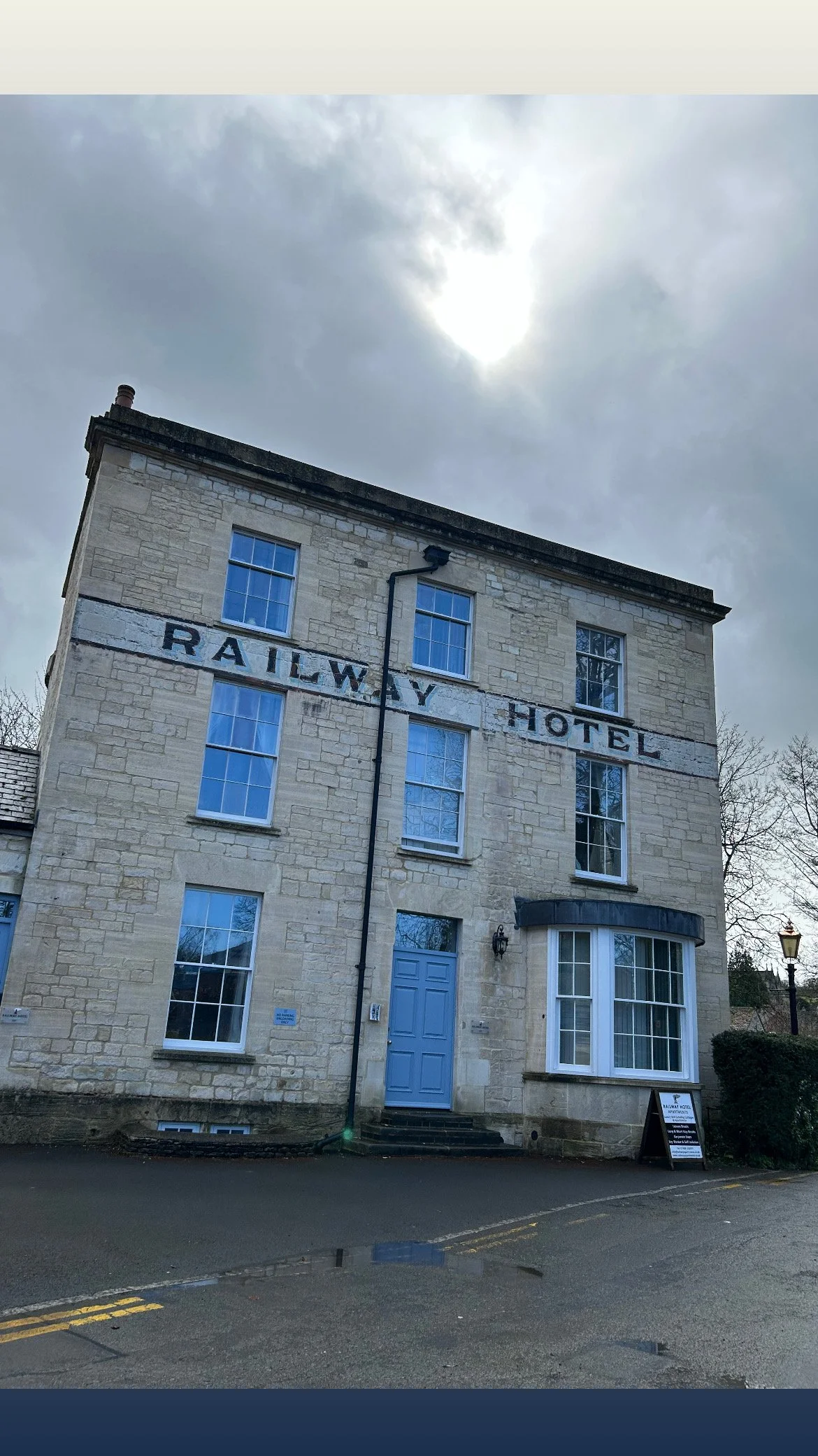 Front view of an old stone building with a sign reading 'Railway Hotel' painted on the facade. Blue door, several windows, and a small handwritten sign outside. Overcast sky in the background.