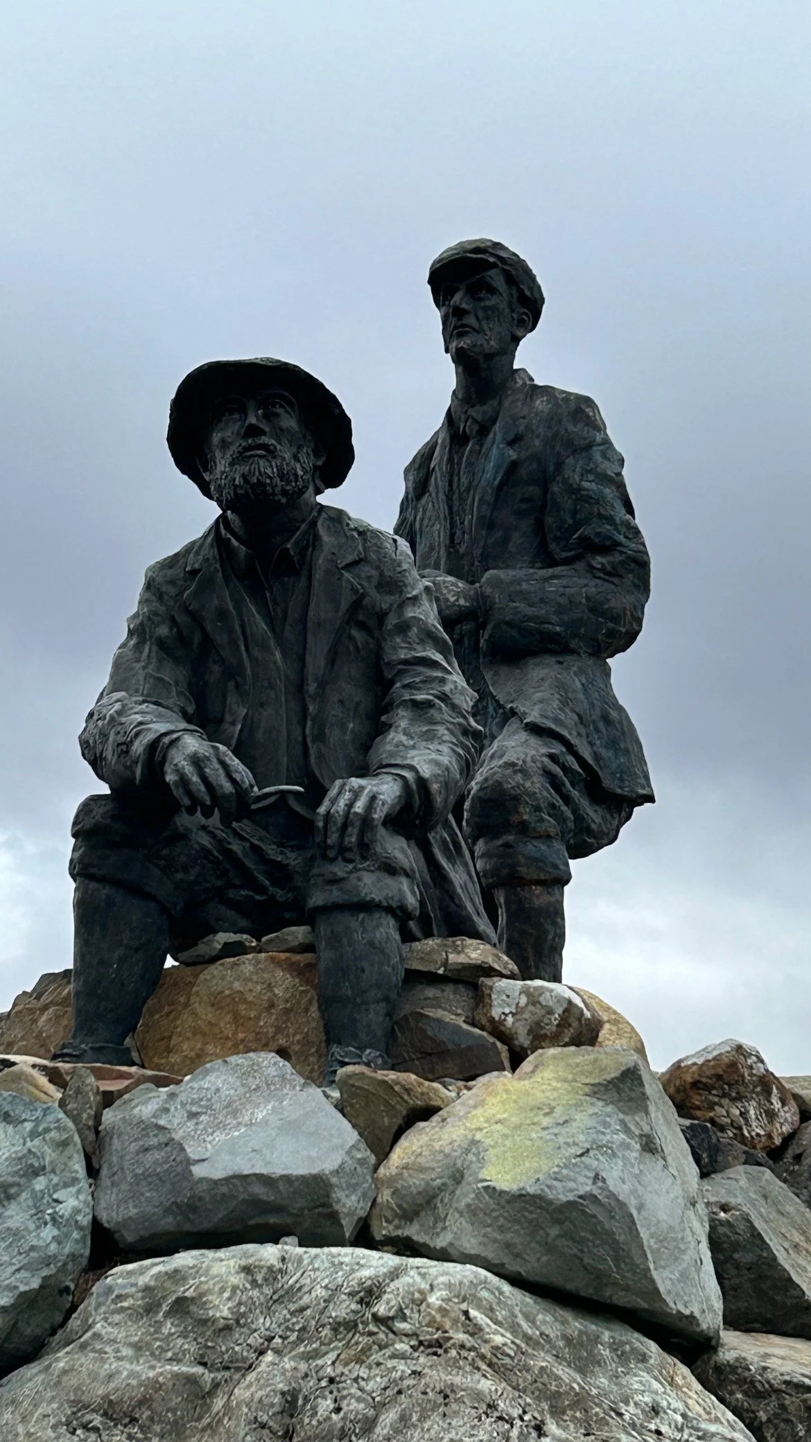 Bronze statue of two men, one seated and one standing, with rocky terrain and cloudy sky in the background.