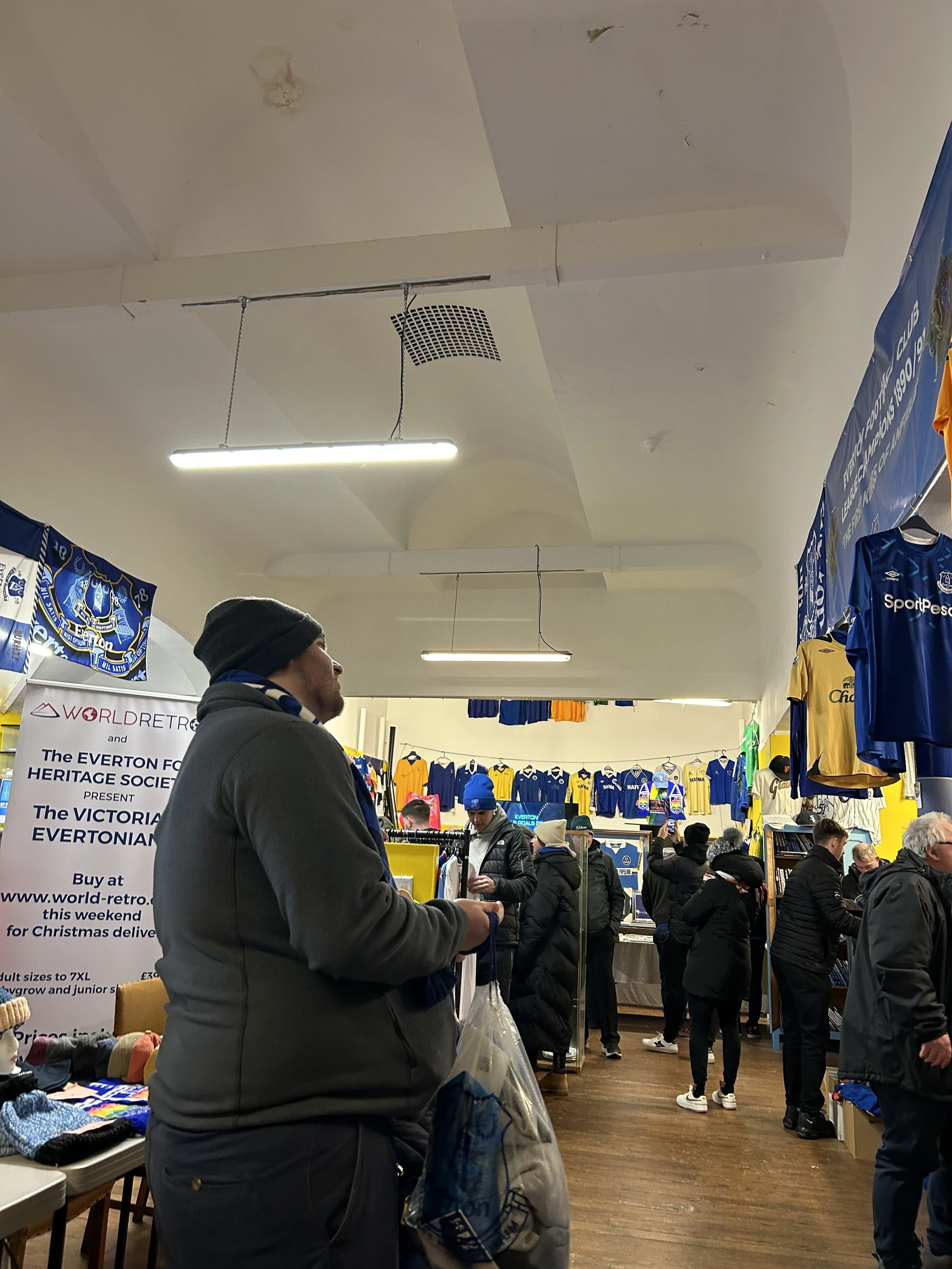 A crowded indoor market or store selling Everton football club memorabilia, including jerseys and scarves, with shoppers browsing and purchasing items.