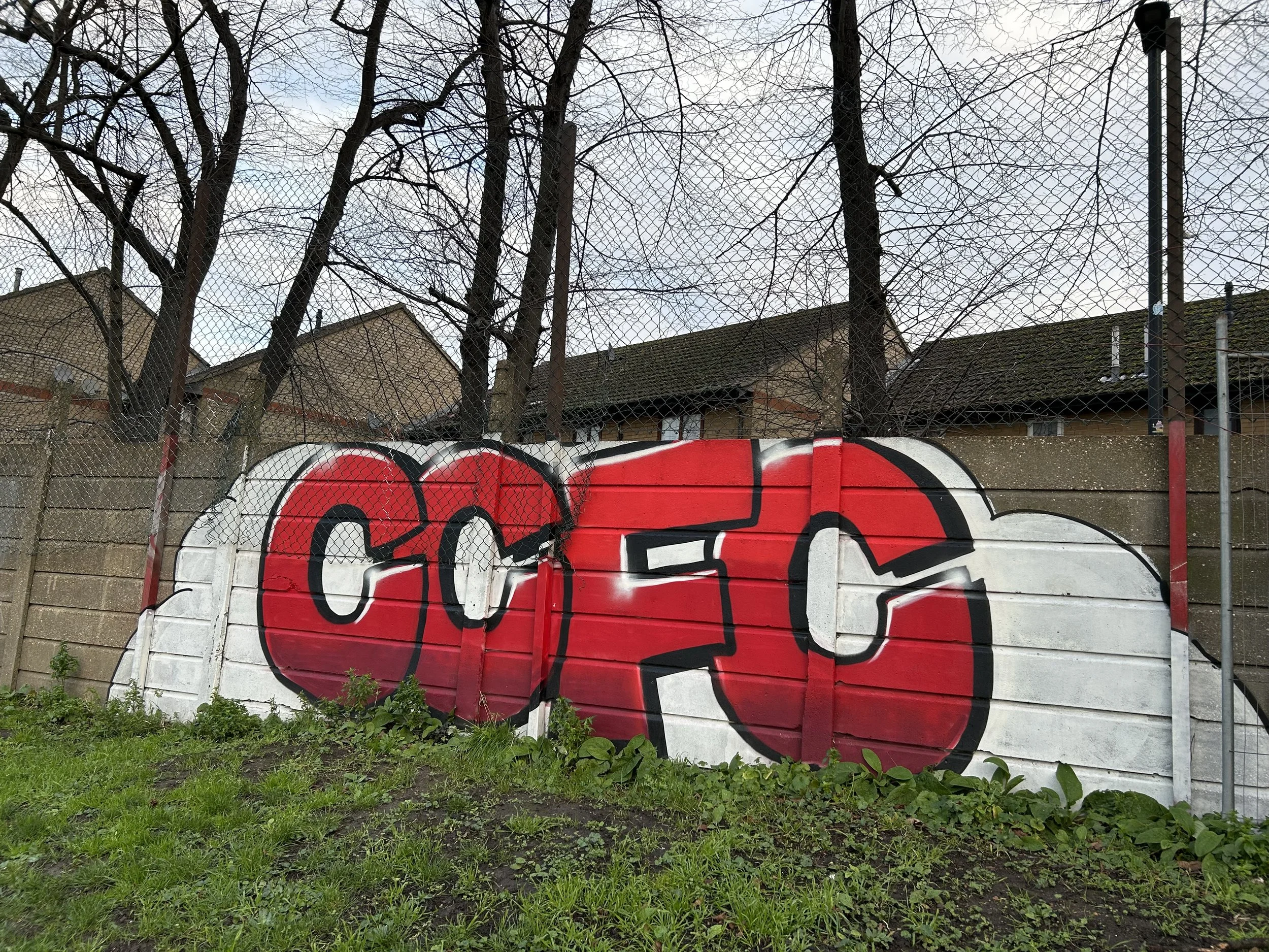 Graffiti artwork with the word 'CCFC' painted in bold red and white colors on a concrete wall next to a chain-link fence, with trees and houses in the background.