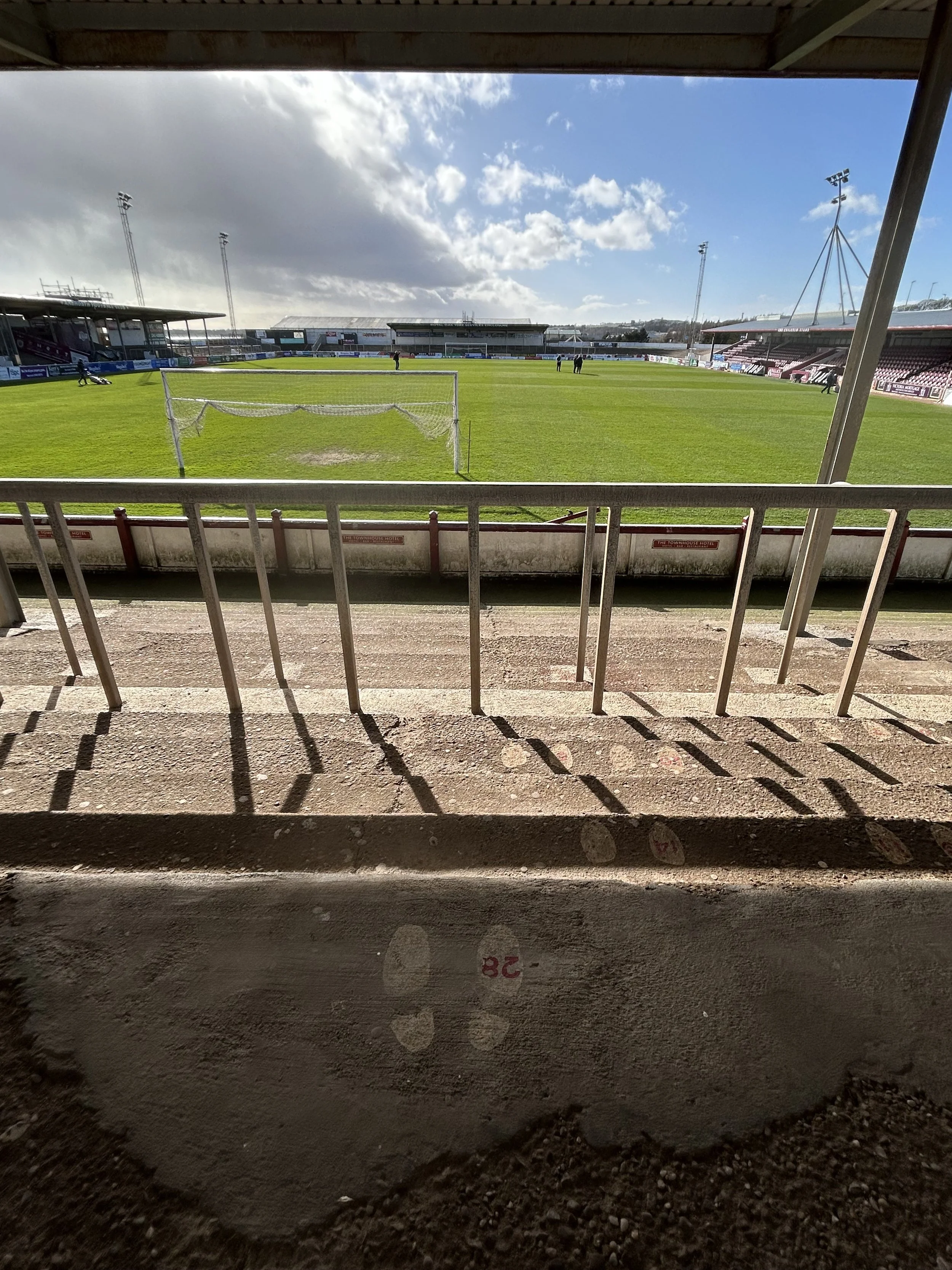 View of a soccer stadium field seen from the stands, with green grass, a goalpost, and a few people in the distance under partly cloudy skies.