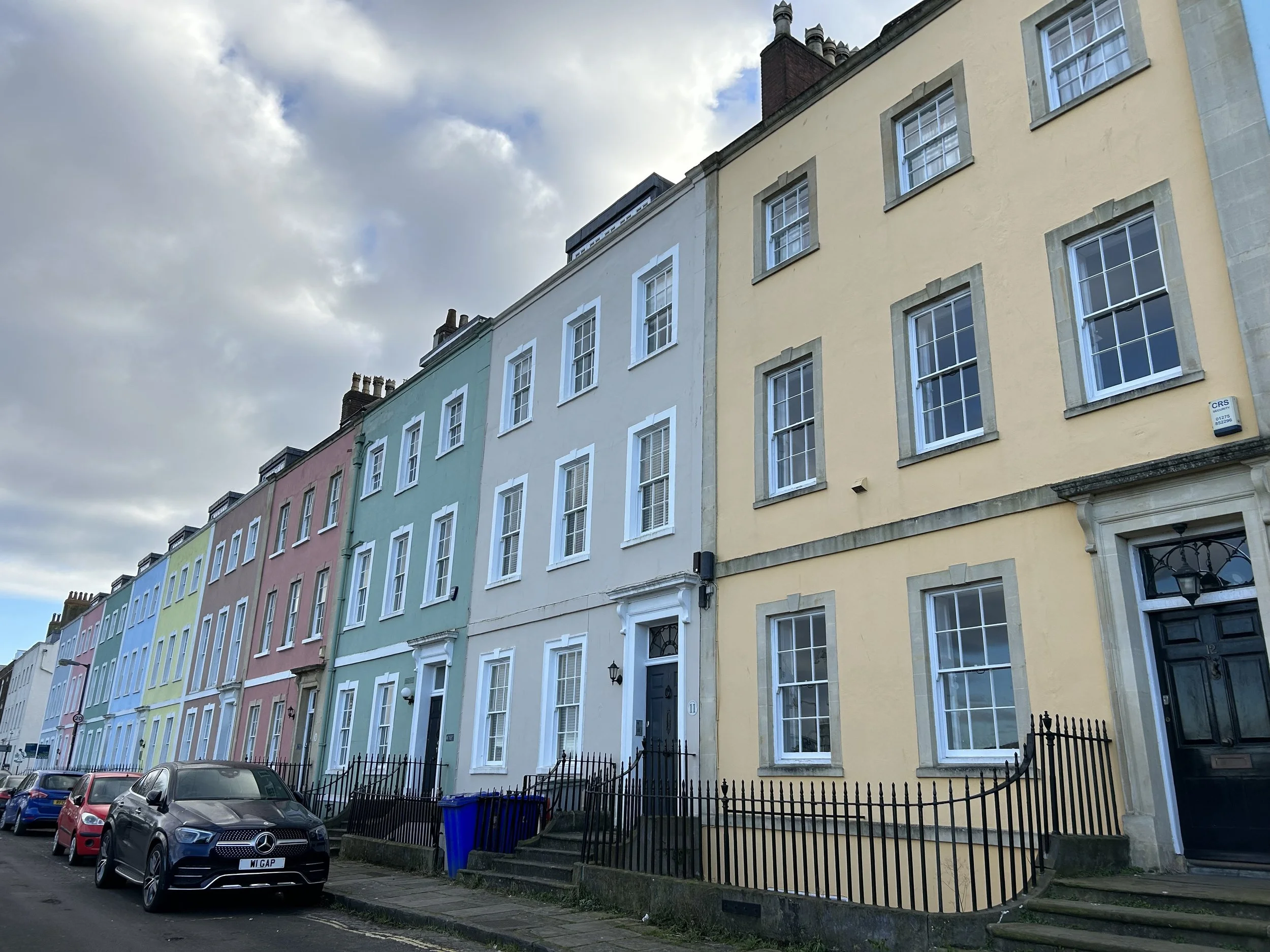 A row of colorful townhouses with pastel facades, black railings, and parked cars on the street under a cloudy sky.