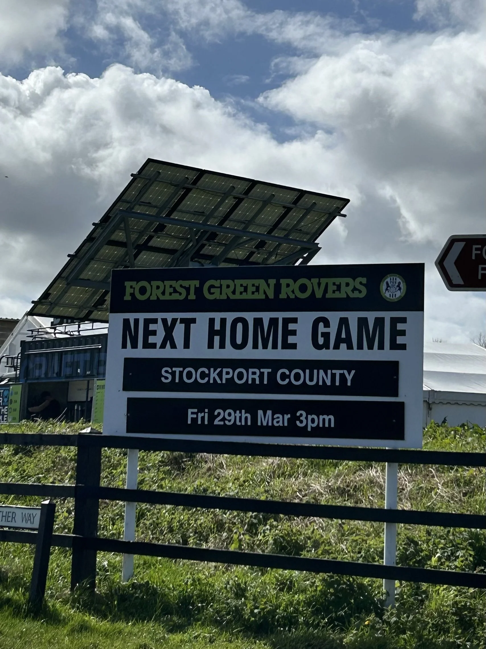 A sign advertising the next home game for Forest Green Rovers against Stockport County on Friday, March 29th at 3 pm. The sign is outdoors with a cloudy sky, and solar panels are visible above it.