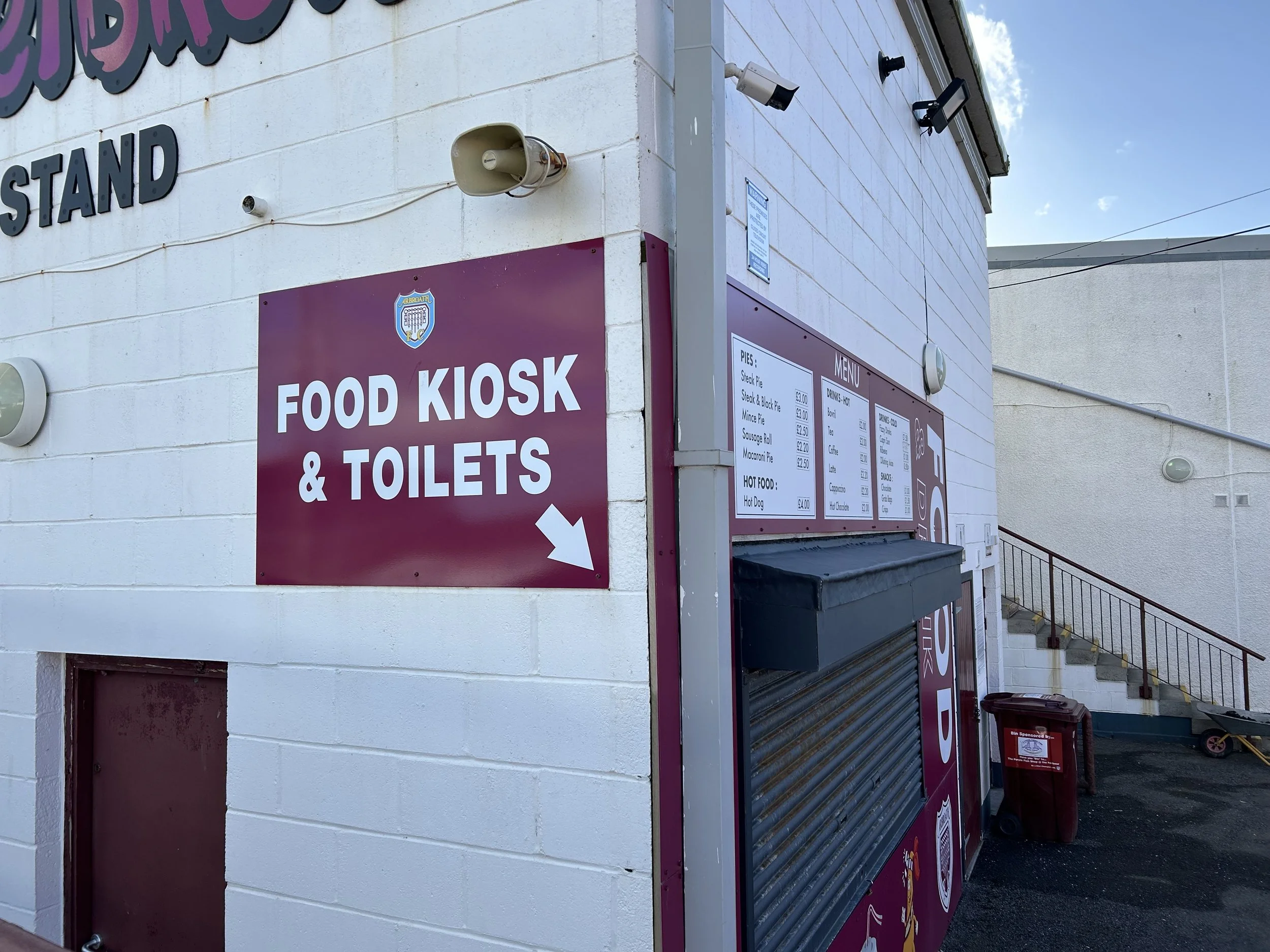 A food kiosk offering food and toilet facilities, located in an outdoor area with stairs and a trash bin nearby.