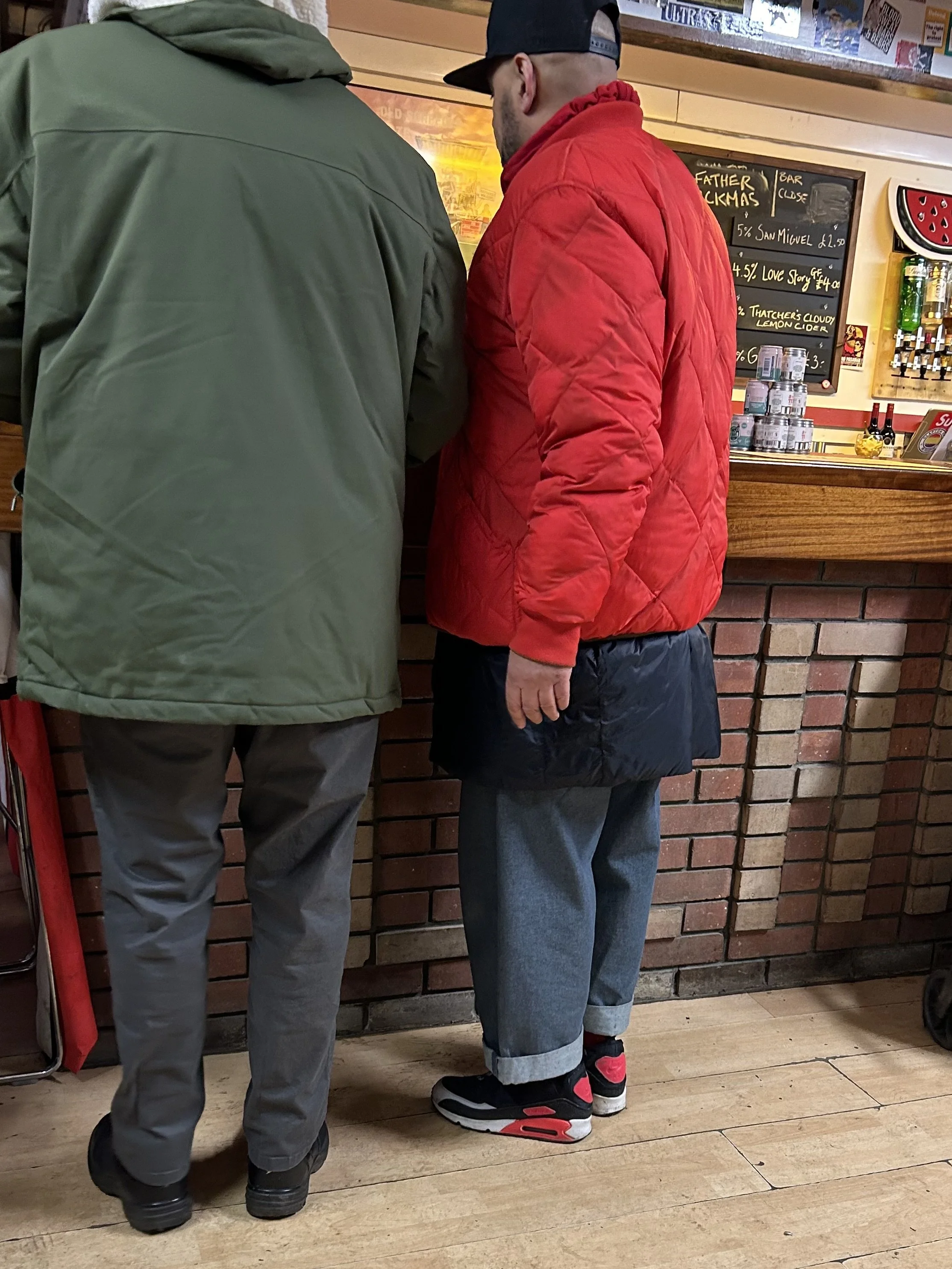 Two men standing at a bar counter, seen from the back. One wears a green jacket and gray pants, the other wears a red jacket, gray pants with rolled cuffs, and black and red sneakers. The bar has a brick wall and a chalkboard menu behind them.