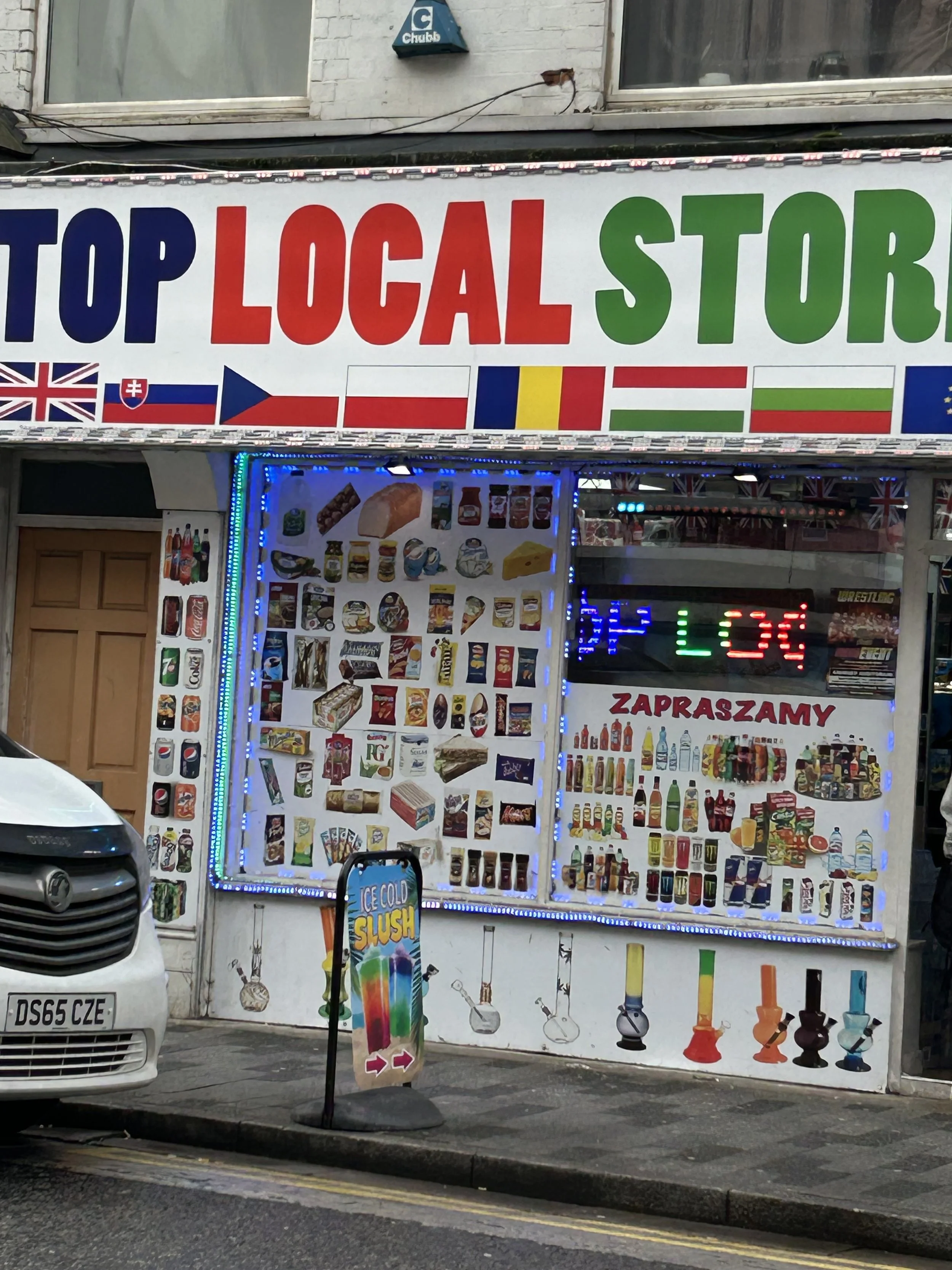 Street view of a small Shop with signs indicating it sells American, English, Slovak, Polish, Romanian, Bulgarian, and European products. The shop window displays various packaged foods, drinks, and smoking accessories, including colorful bongs and a