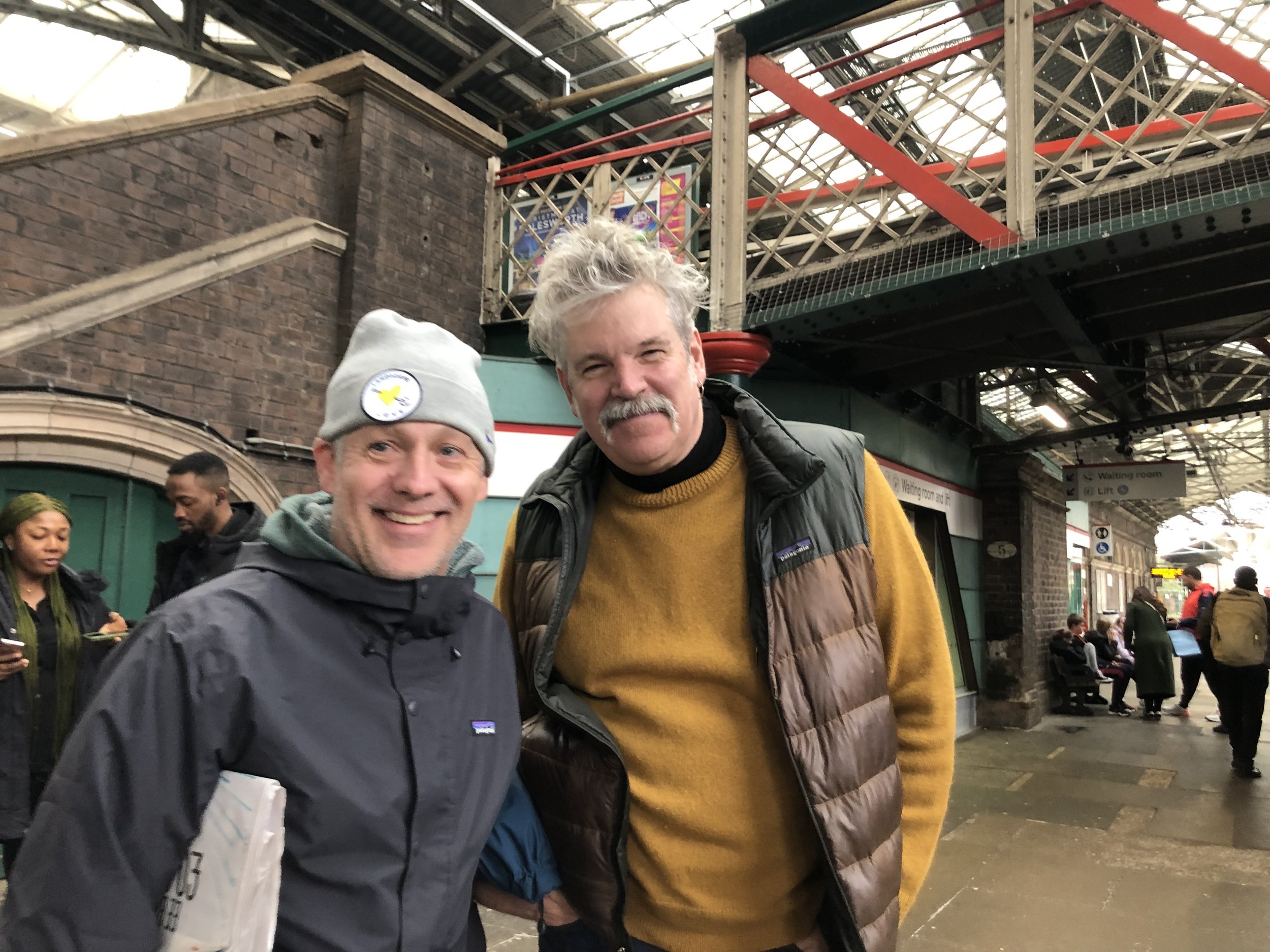 Two smiling men standing at a train station, with other people sitting and walking in the background. The man on the left wears a gray beanie with a yellow patch and a gray jacket. The man on the right has gray hair and a mustache, wearing a yellow s