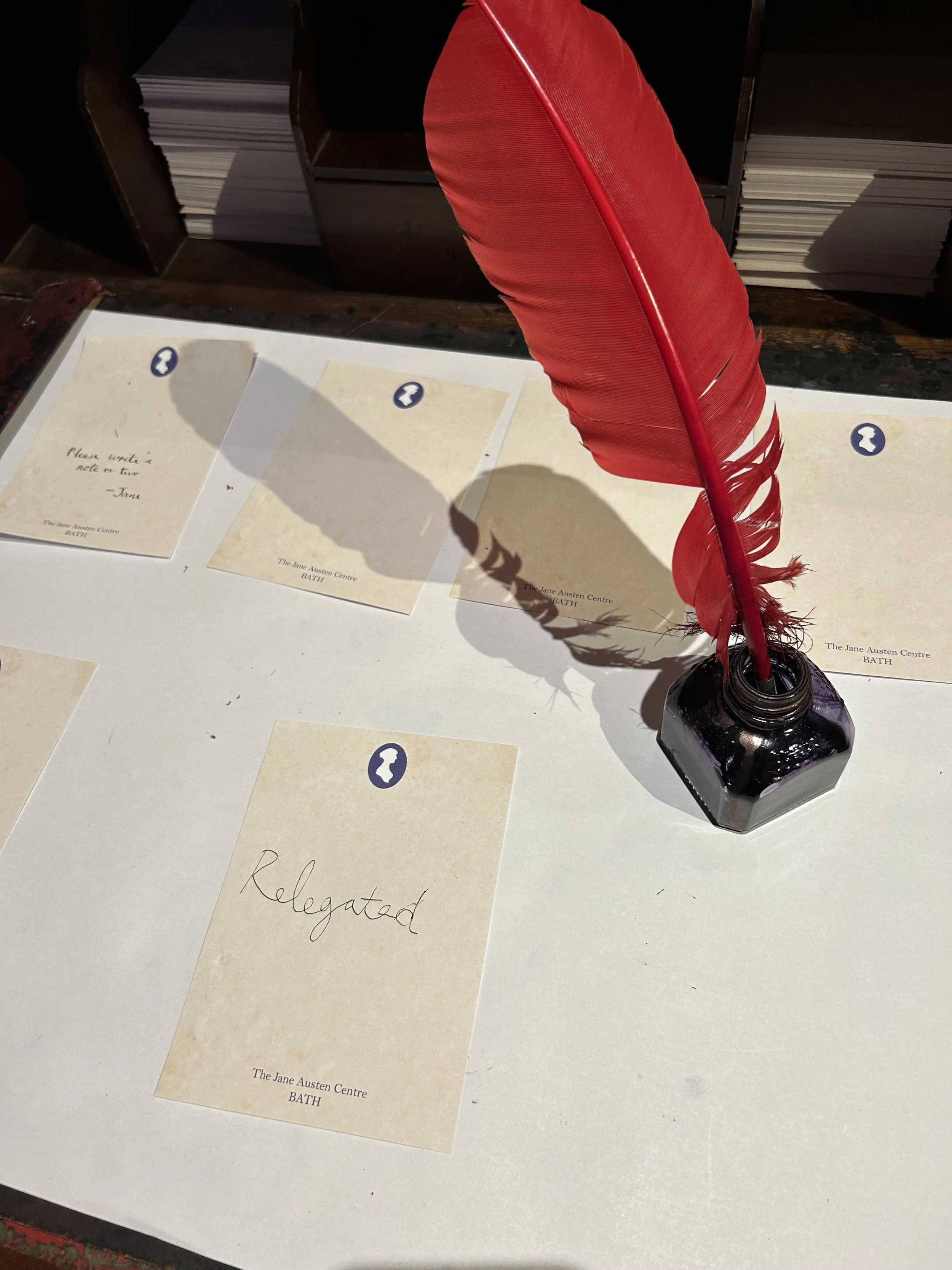 A red feather quill pen in an ink bottle on a table with handwritten notes and printed cards, with shelves of books or files in the background.