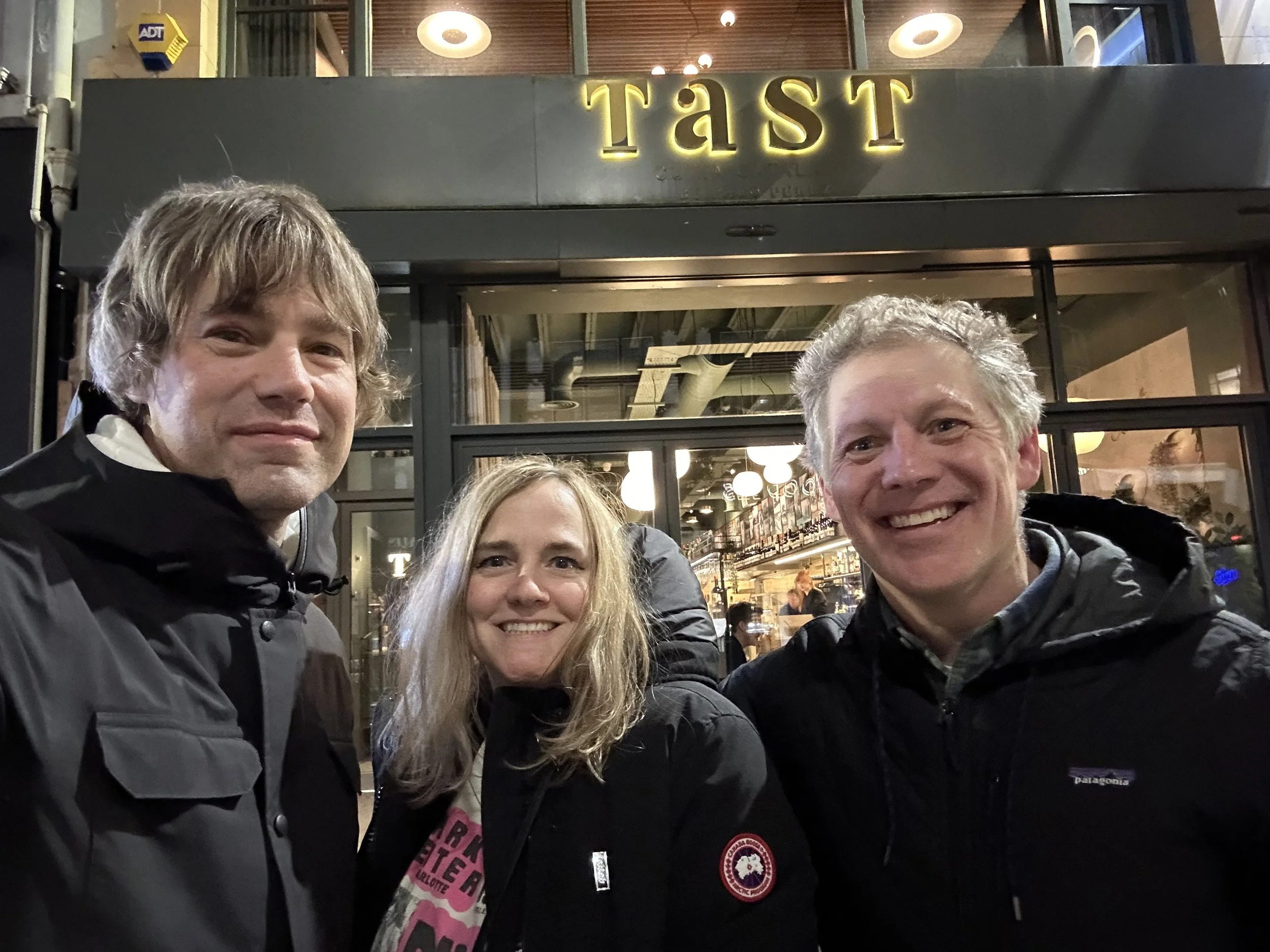 Three smiling adults wearing black jackets are standing in front of a restaurant with a sign that says 'TÄST' at night.