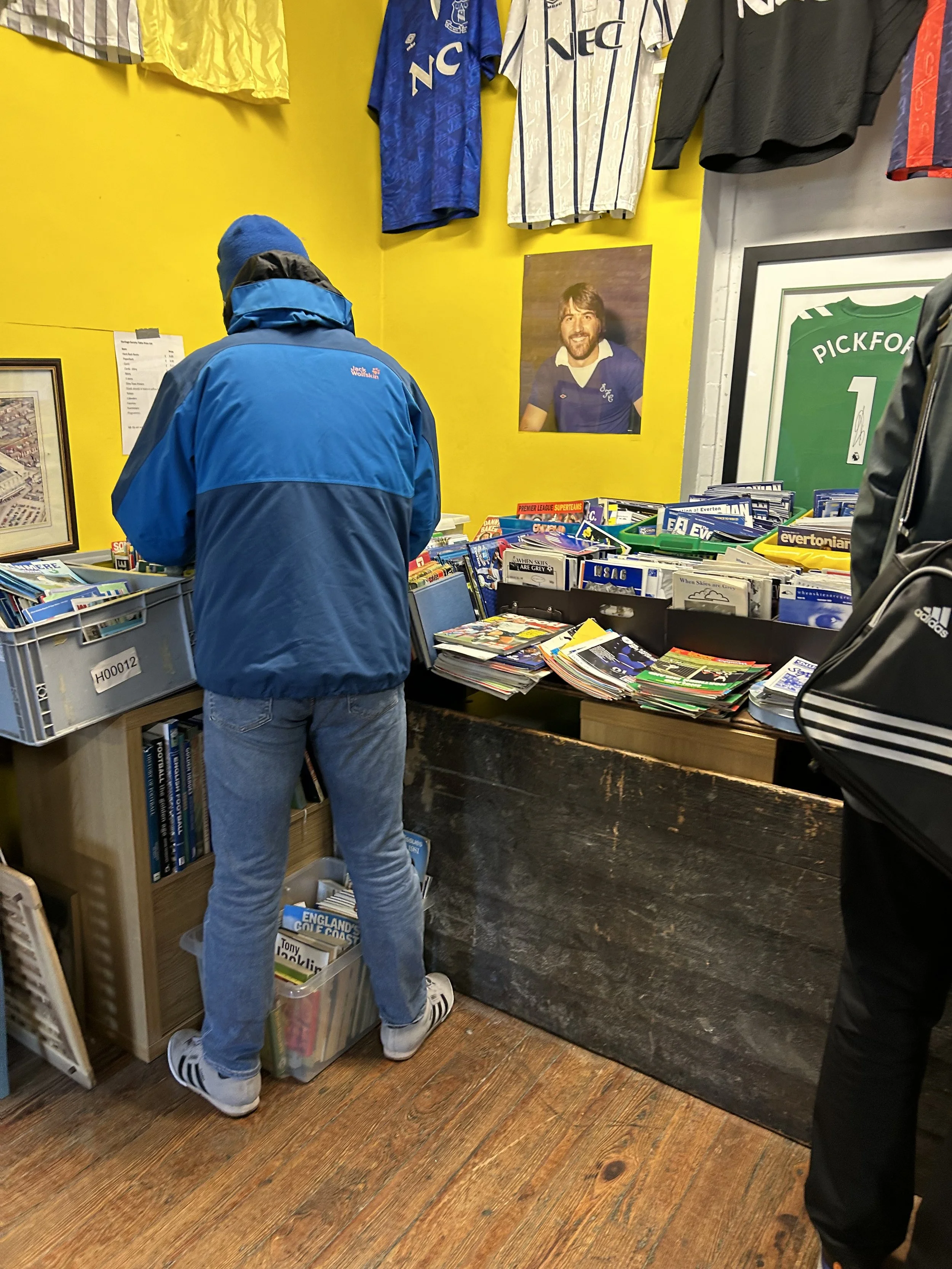 Person wearing a blue jacket and jeans browsing books or magazines at a stand inside a store with sports memorabilia, including framed jerseys, a poster of a man, and a framed green jersey on the wall.