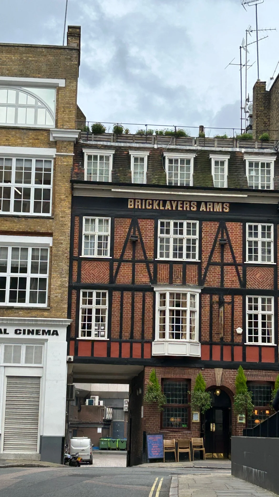 A street view featuring a historic brick building with a black timber frame facade named 'Bricklayers Arms.' The building has multiple windows, some with white frames, and a small bay window projecting from the front. Adjacent to it is a modern white