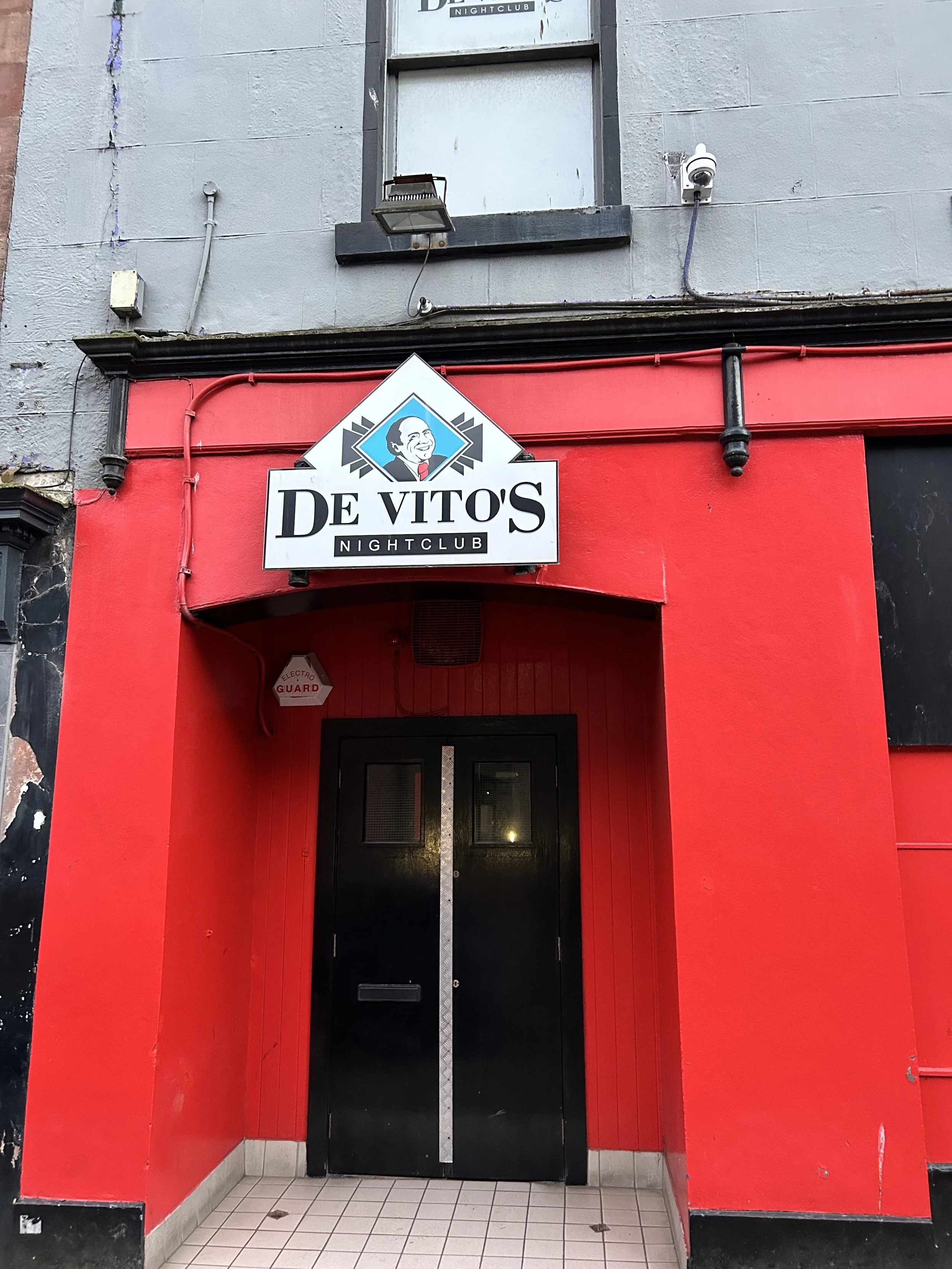 Entrance to De Vito's Nightclub with red and black exterior, black double doors, and a sign displaying a smiling man in a suit along with the club's name.