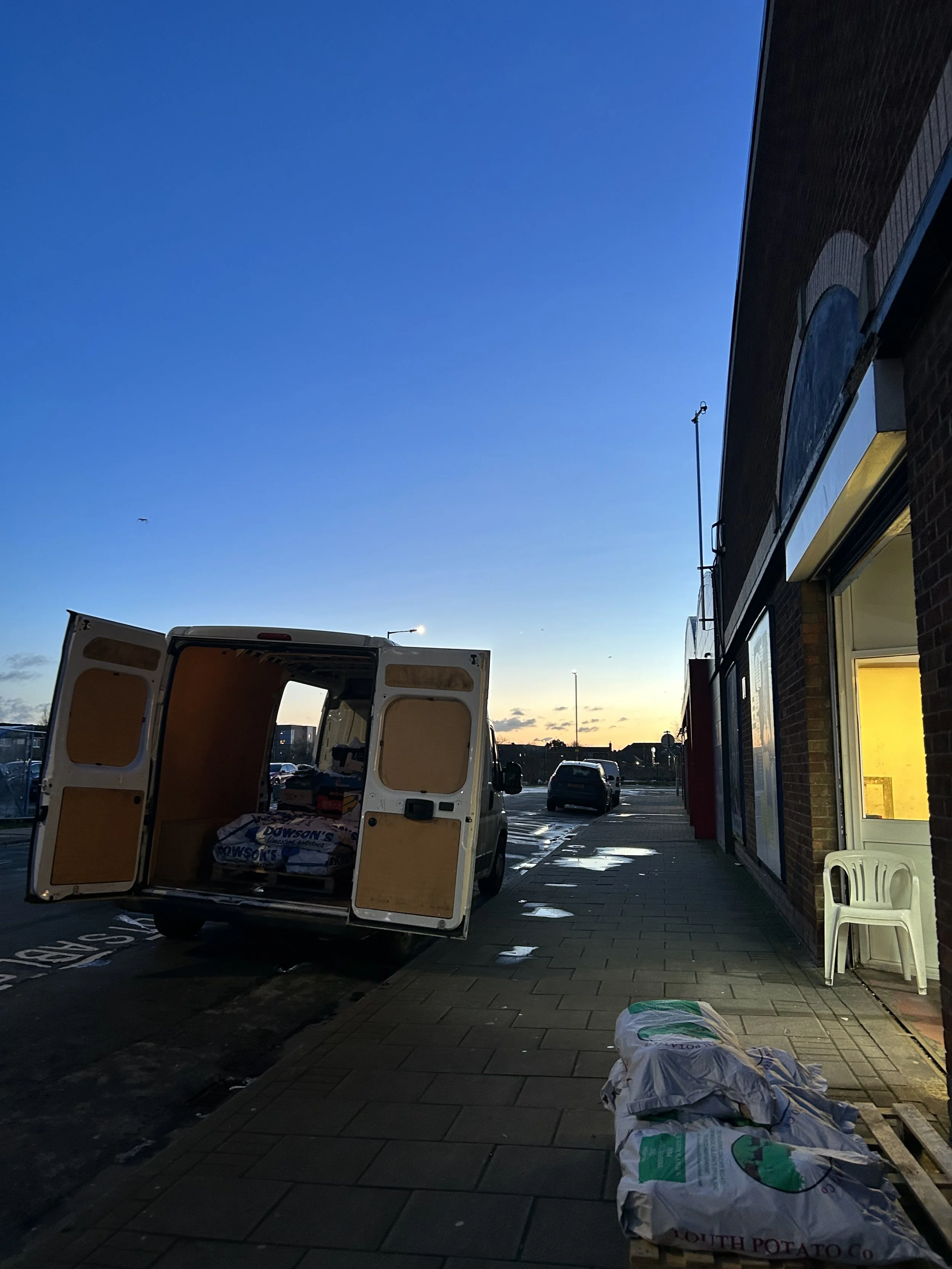 A parking lot at dusk with a white delivery van parked, its rear doors open revealing packages inside. A few bags are on the sidewalk near the van. The building on the right has a window lit from inside and a stack of white plastic chairs outside. Th