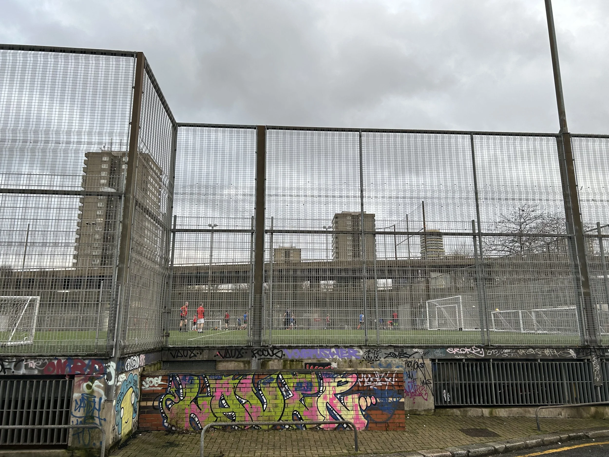 Fence surrounding an outdoor soccer field with players practicing, graffiti on the wall below, high-rise buildings and an overcast sky in the background.