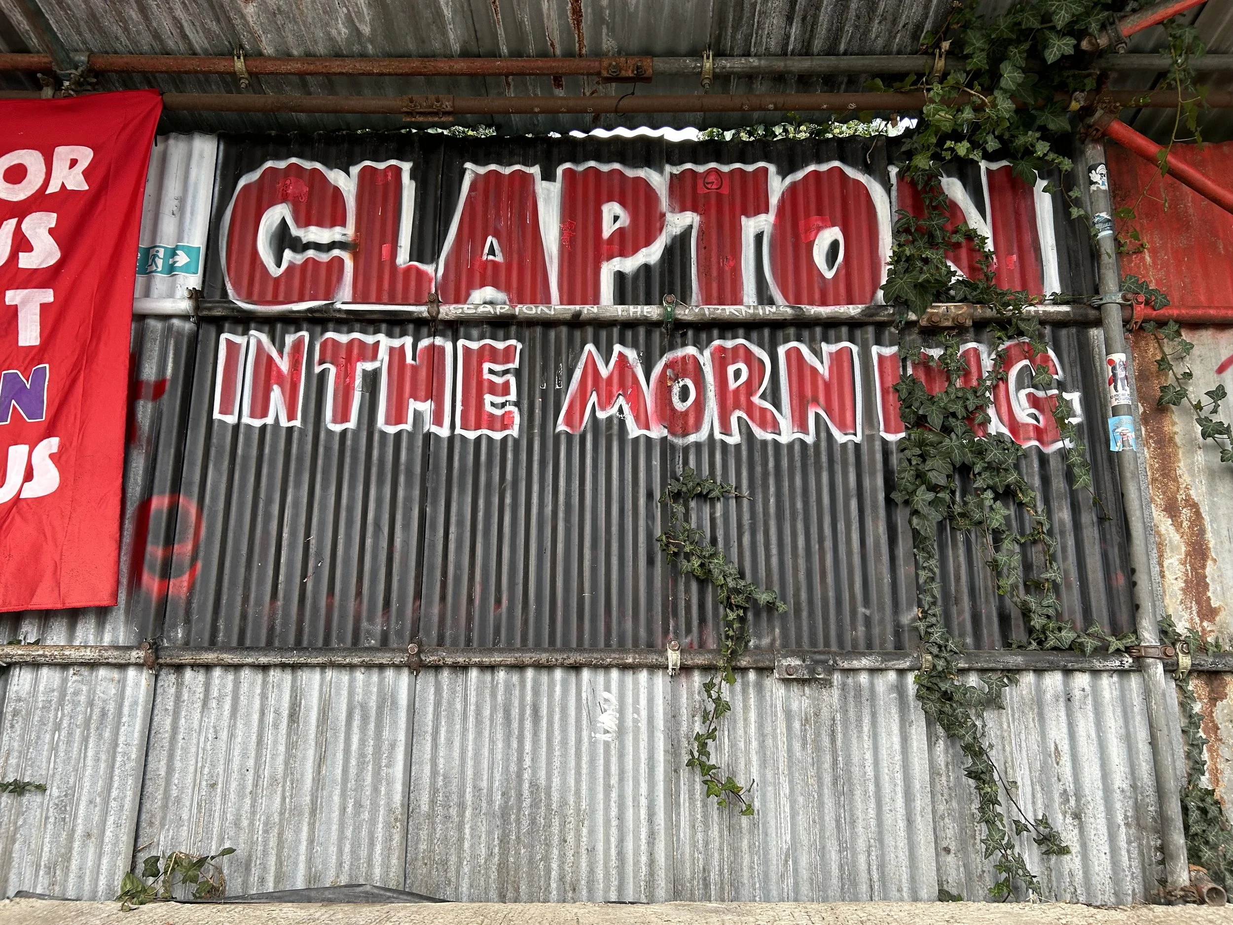A sign made of corrugated metal with painted red and white letters reading 'CLAPTON IN THE MORNING.' There are some green ivy plants growing on the right side of the sign.