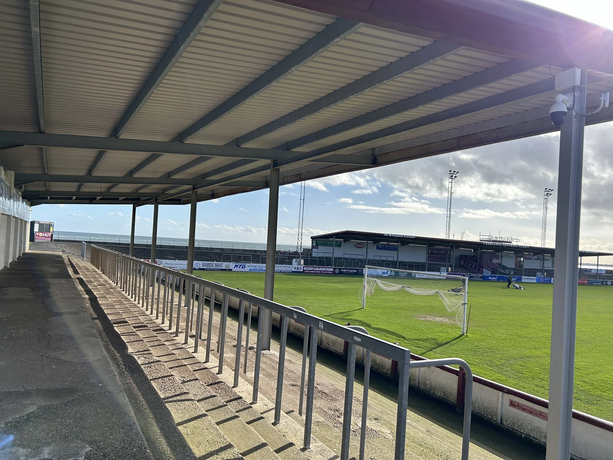 Empty sports stadium with a green field, a soccer goal net, and a covered seating area with metal railings on a sunny day.