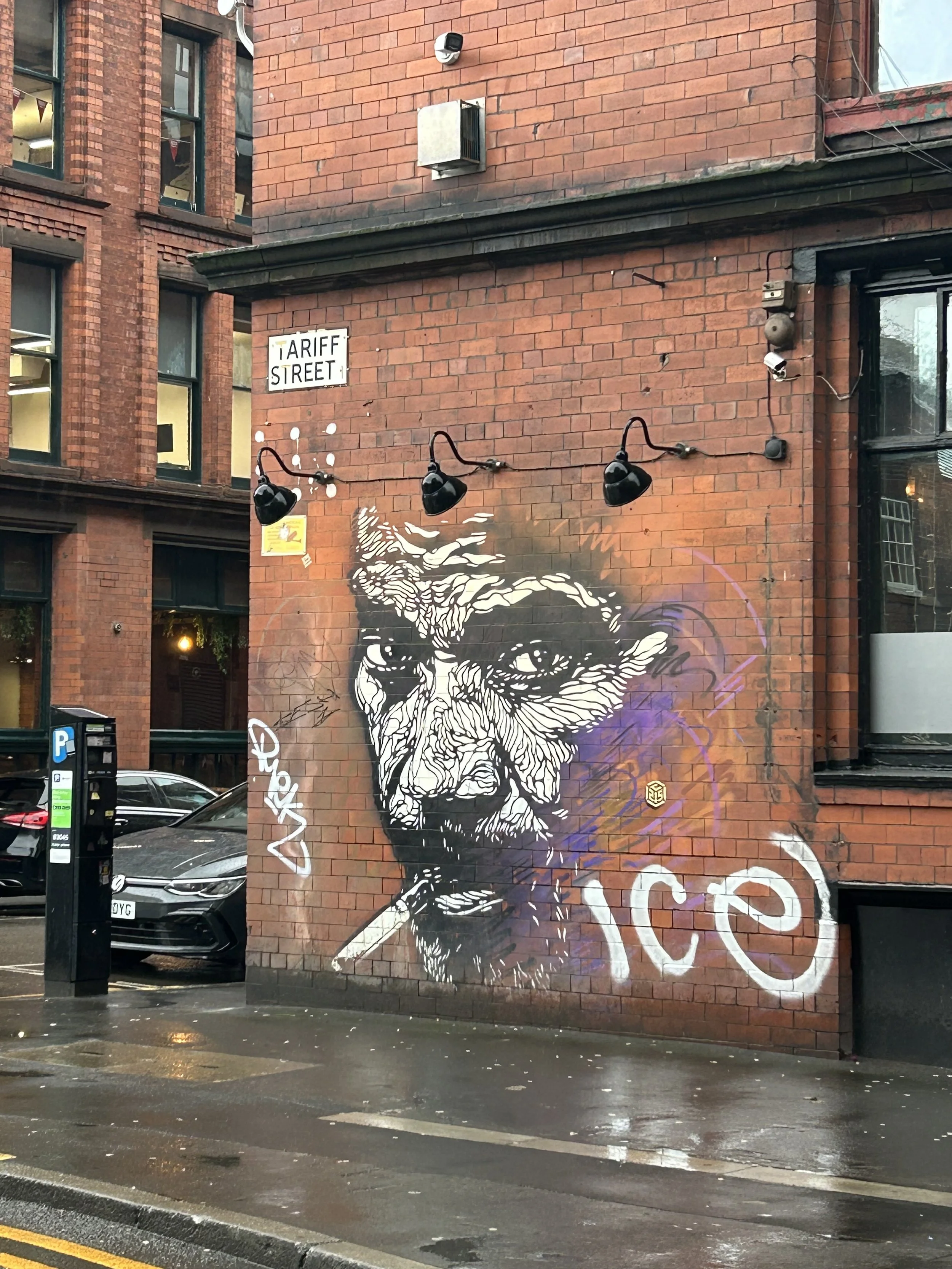 Street art mural of a man's face with intense expression, painted on a red brick wall. The mural is black and white with some purple graffiti tags, and a cigarette in the man's mouth.