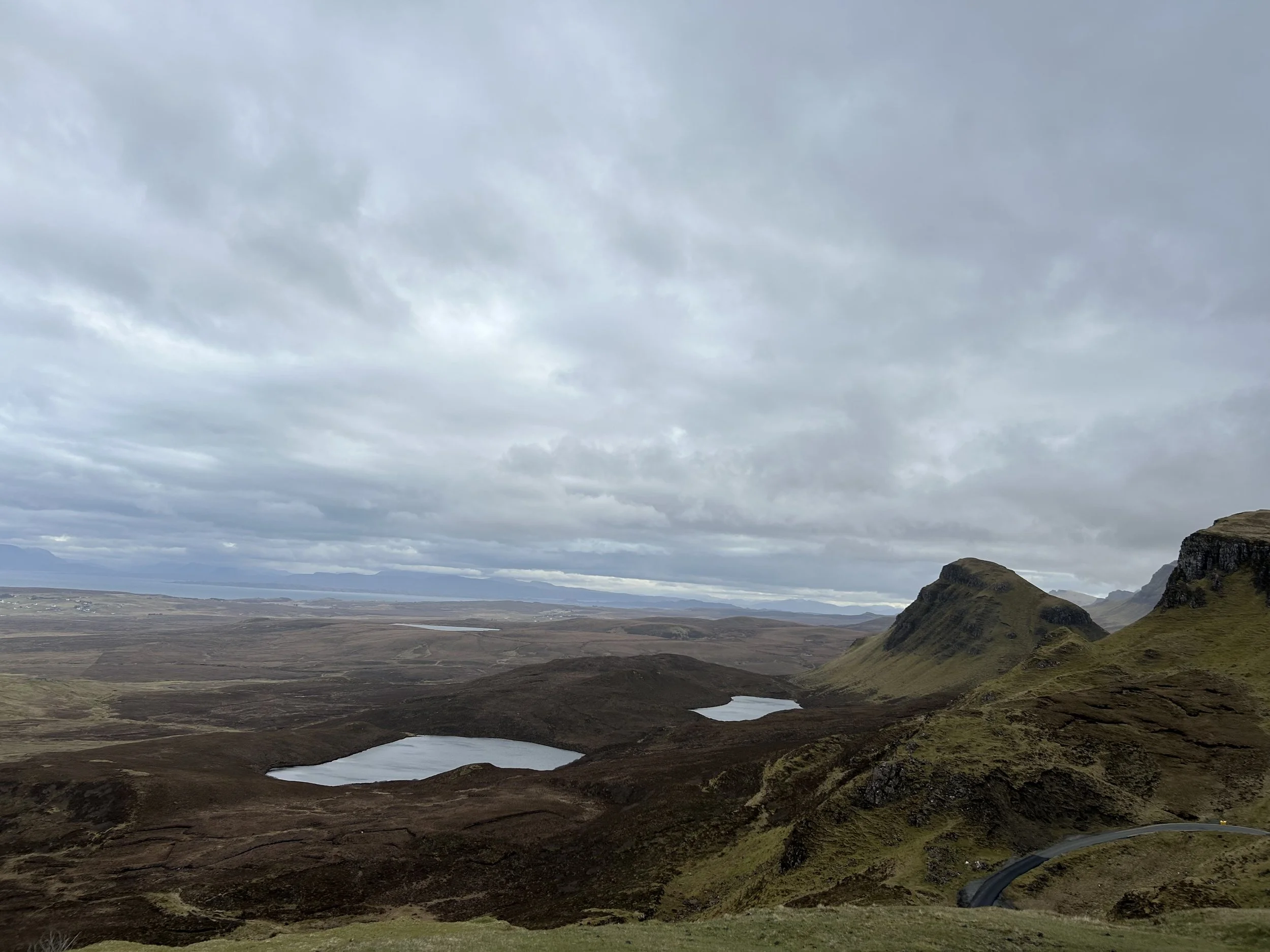 A vast, cloudy landscape with rolling hills and small lakes, possibly in Iceland, with green grass and mountains in the distance.