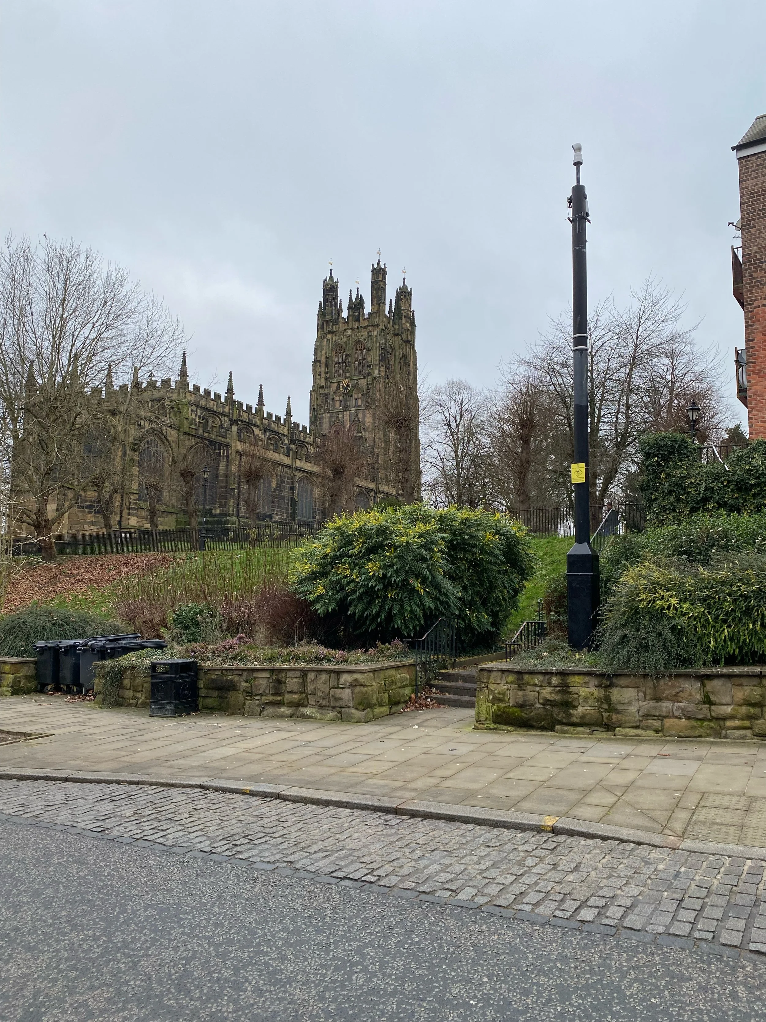 A gothic-style church with a tall, ornate tower, surrounded by trees and bushes, with a paved sidewalk, street, and lamppost in the foreground on a cloudy day.