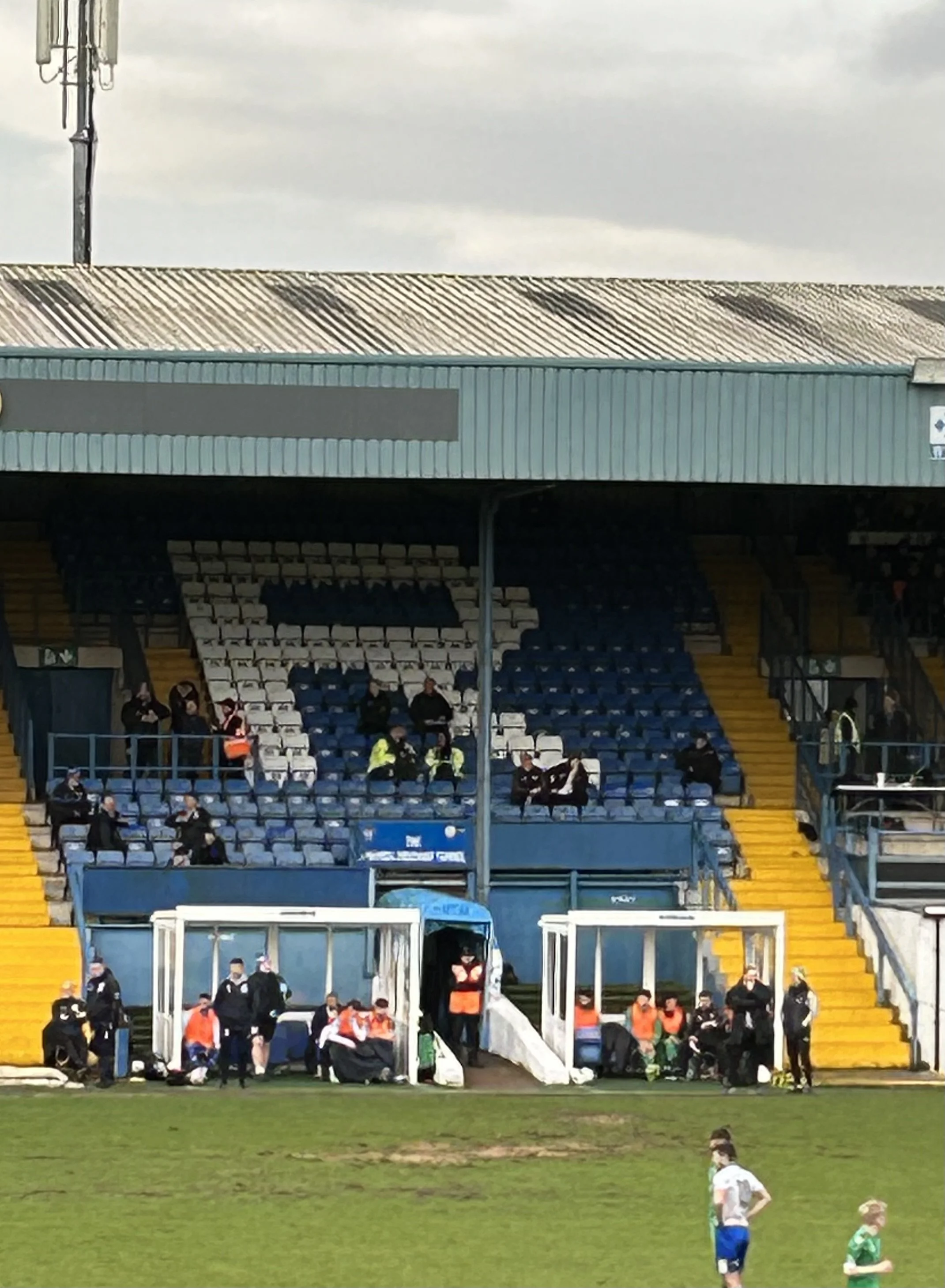 View of a soccer stadium with empty blue and white seats and a few people sitting, some standing near the sideline, and players on the field.