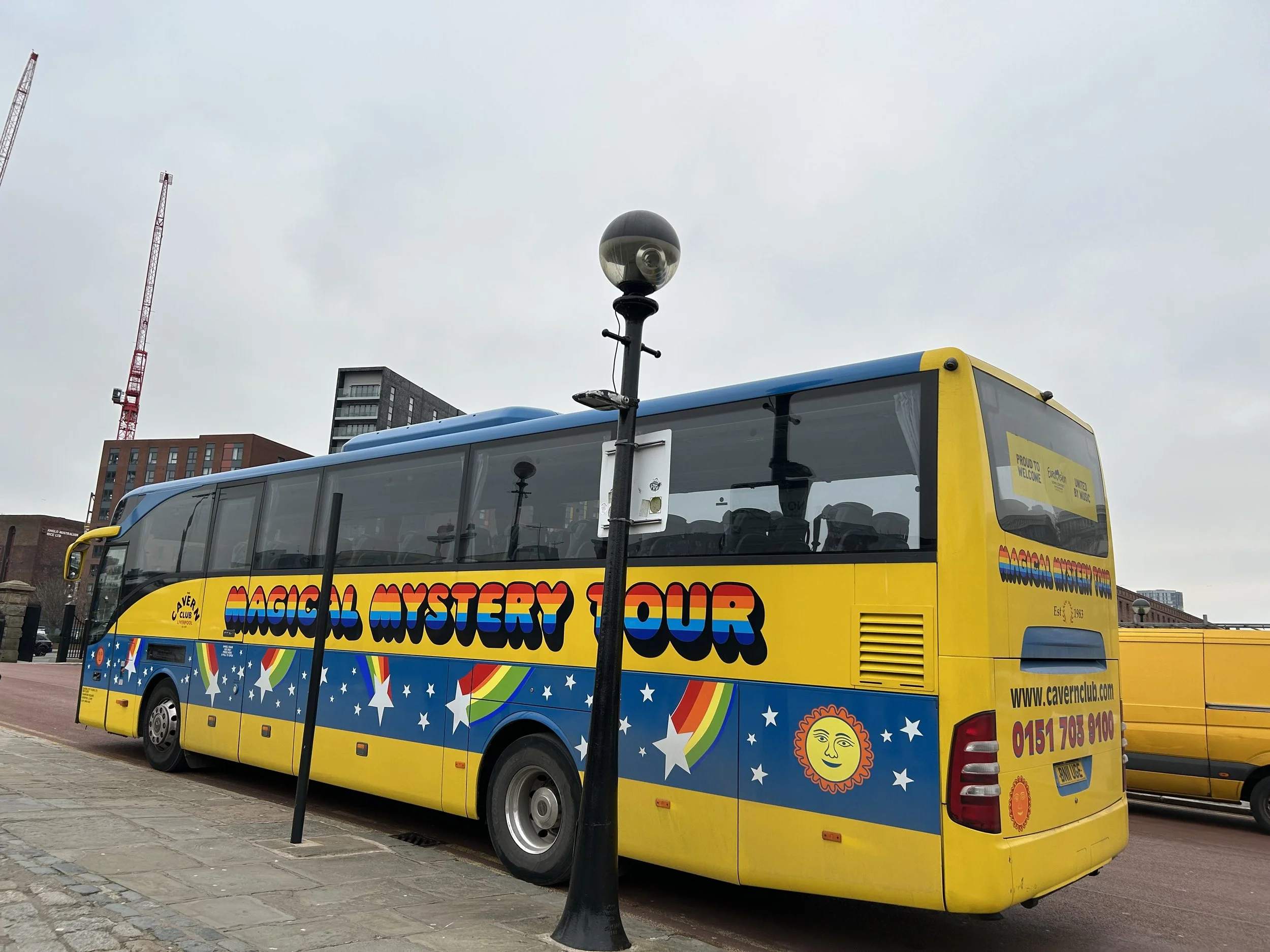 Yellow tour bus with colorful rainbow-themed graphics and the words "Magical Mystery Tour" parked on a city street under a cloudy sky.