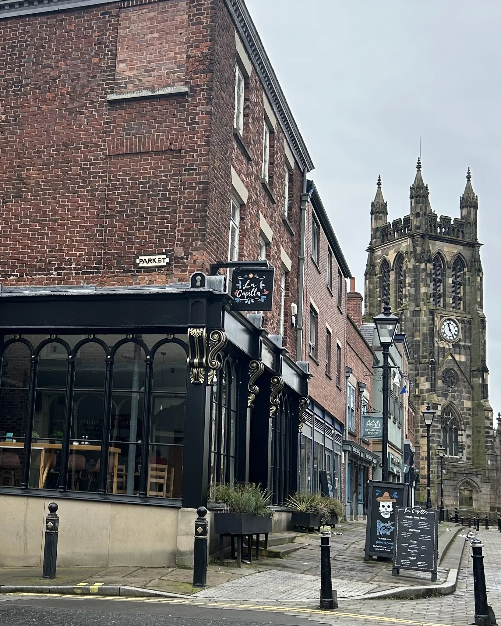 Street view of a historic church with a clock tower and Gothic architecture, nearby buildings with signs for a restaurant and cafes, sidewalk with planters, street lamps, and a chalkboard menu in front of the establishment.