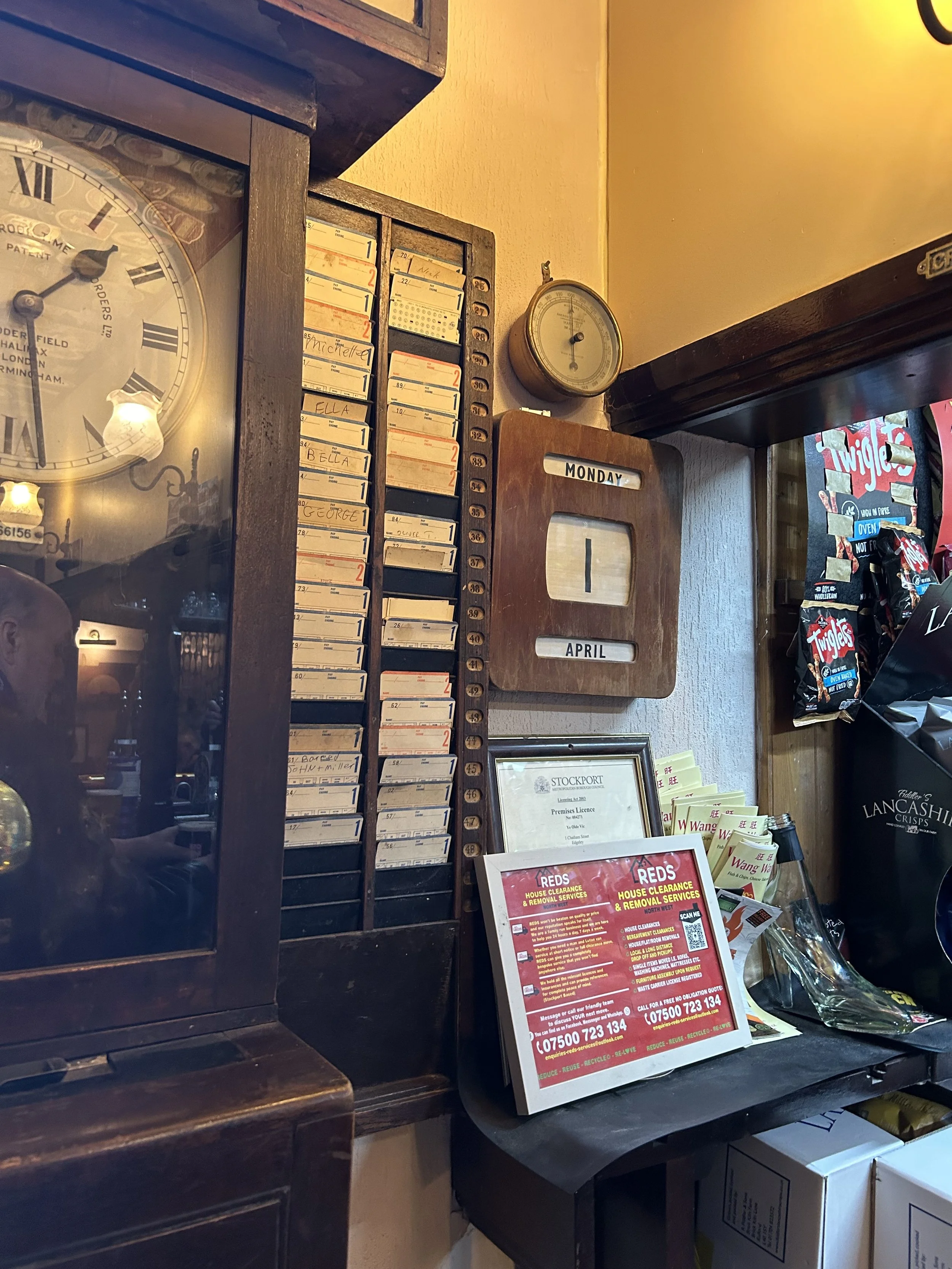 A vintage wall clock, a calendar displaying Monday, April 1, a small round thermometer, an appointment board with names and numbers, and various flyers and snacks on a counter.