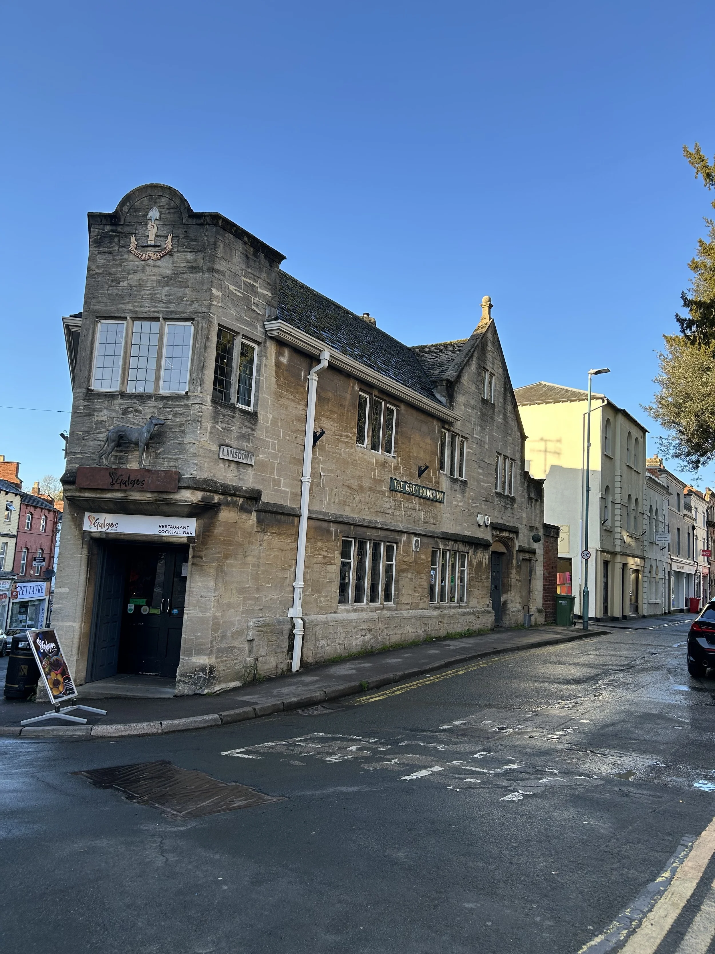 A historic stone building on a street corner with signs for 'The Grey Hound Inn' and 'Galagos Restaurant Cocktails Bar.' The street appears wet from rain, with a clear blue sky above and some other buildings visible in the background.