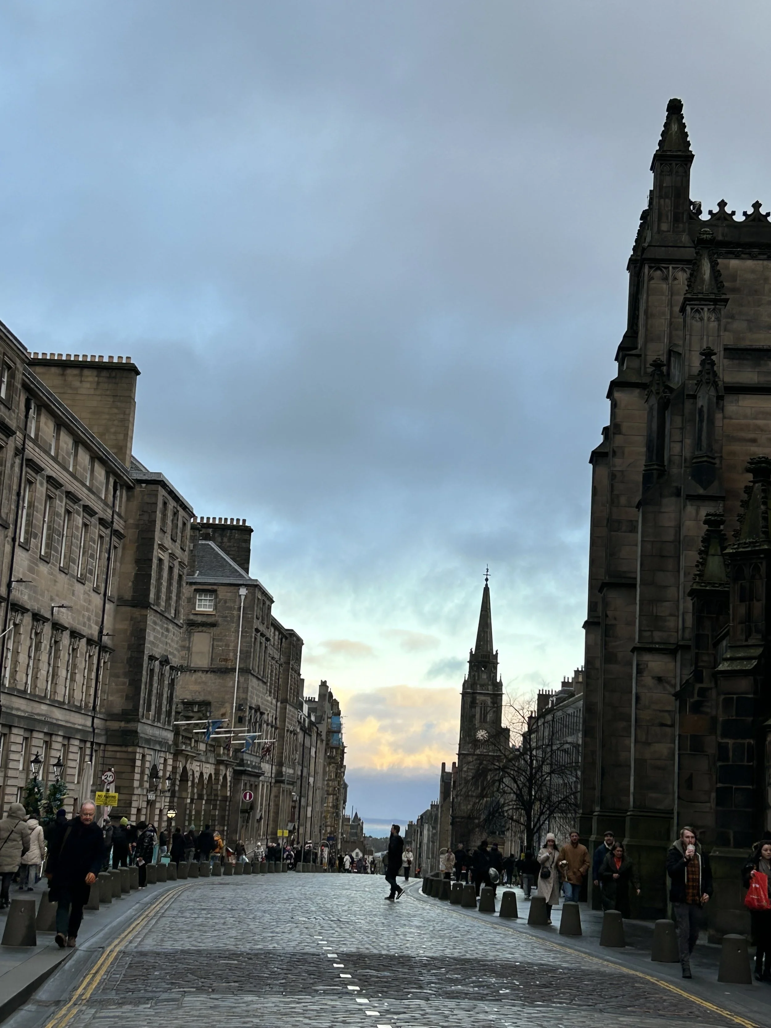 Street scene in a historic city, with cobblestone roads, old stone buildings, and a tall church steeple in the background, under a partly cloudy sky.