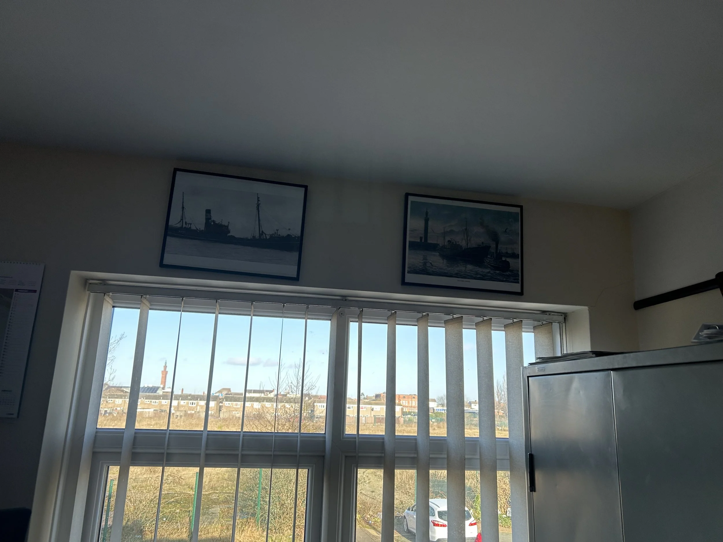 View of a window with vertical blinds, showing a landscape with buildings, trees, and a distant water body. Two framed pictures of ships are hung above the window on the wall.