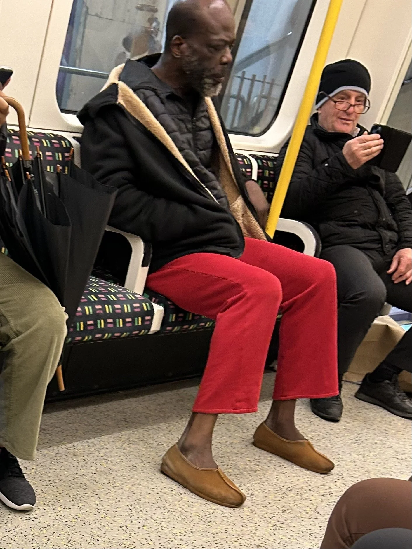 A man with dark skin, grey hair, and a beard wearing a black jacket with a tan hood, red pants, and tan shoes sitting on a subway train next to a man in glasses and a black jacket, who is looking at his phone.