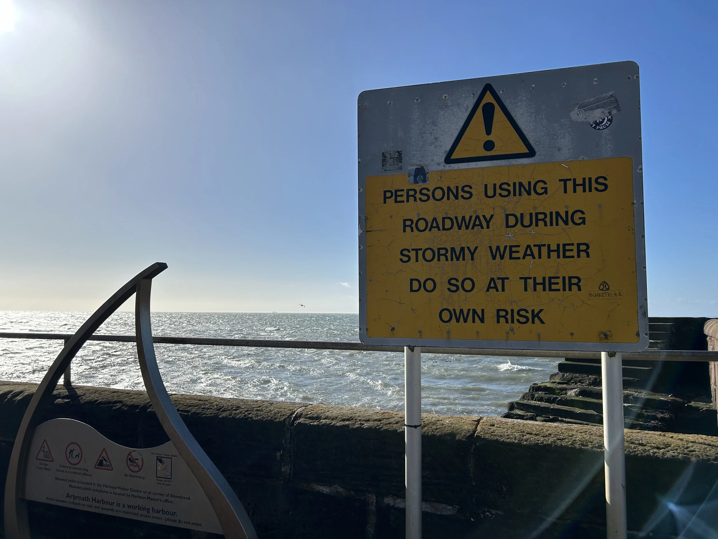 A sign near the ocean warning persons about using the roadway during stormy weather, with the ocean and a seagull in the background.