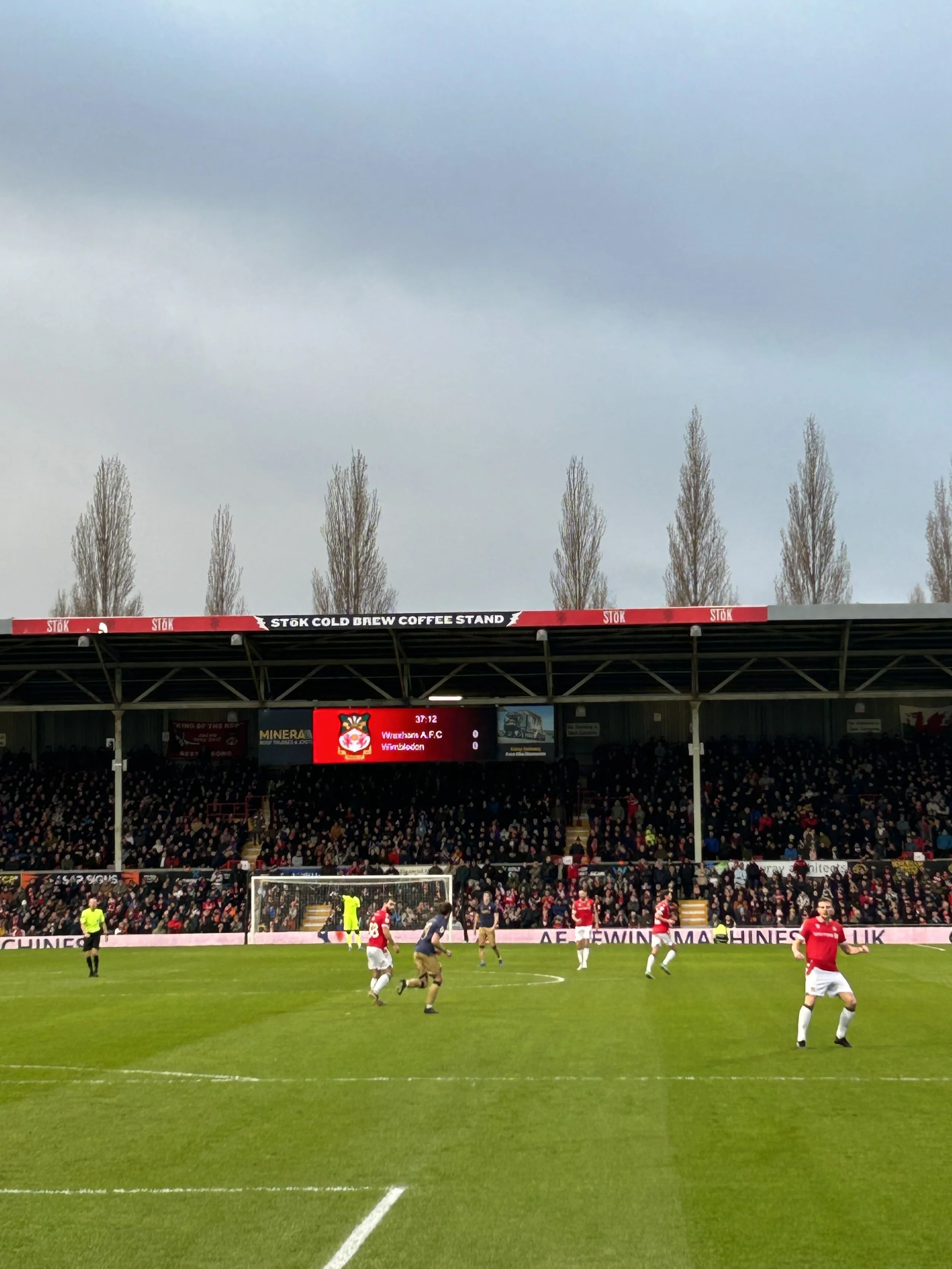 A soccer match in progress at a stadium with players in red and navy uniforms. The stands are filled with spectators, and a scoreboard shows a score of 0-0 for Wenbledon and Warwick A.F.C. The stadium has a sign for a cold brew coffee stand.
