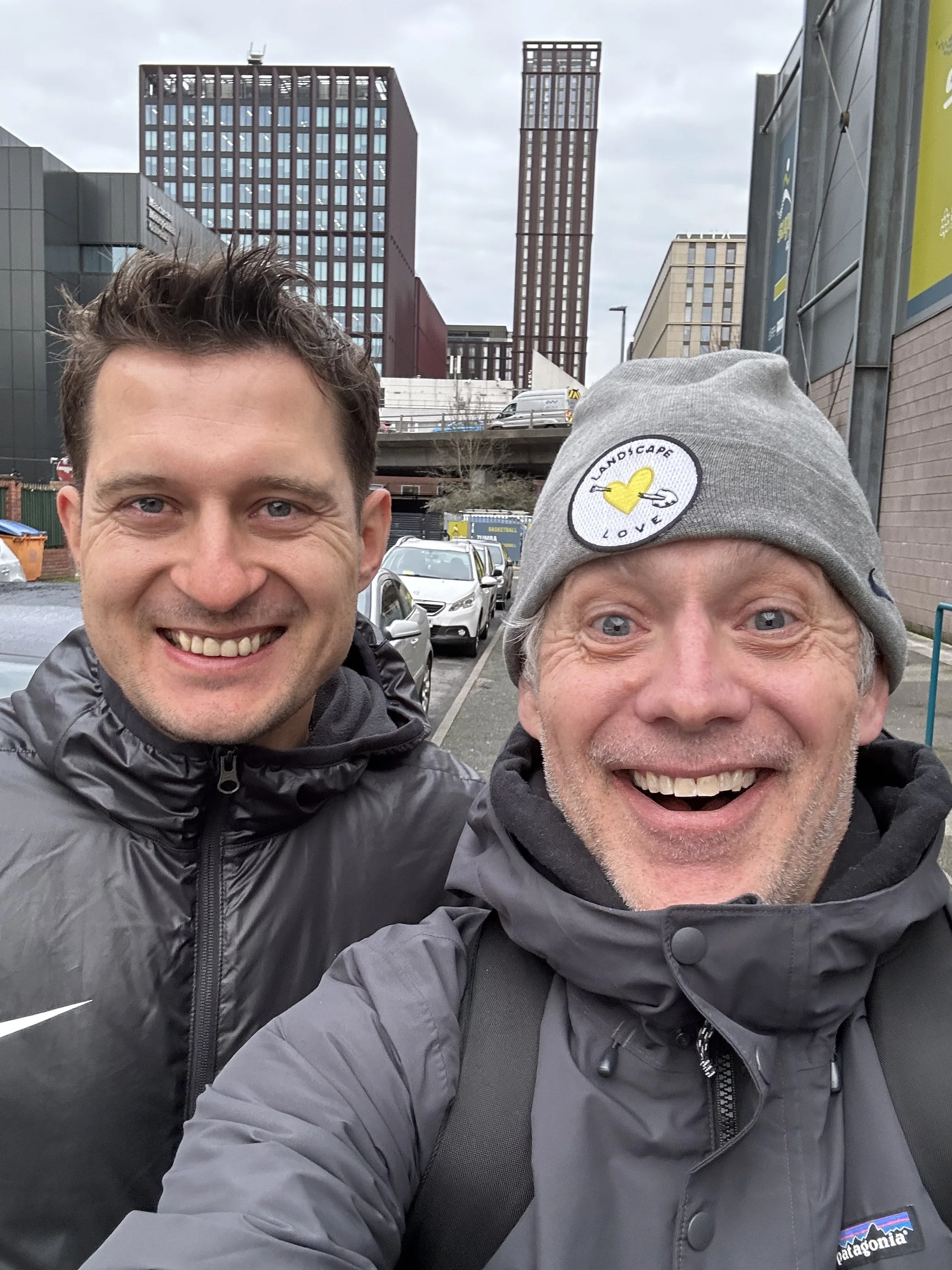 Two smiling men taking a selfie outdoors in an urban area with tall buildings and parked cars in the background.