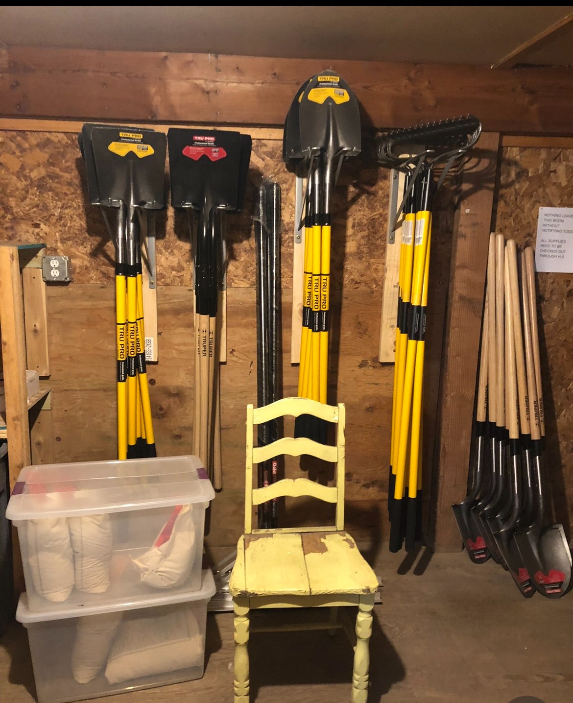 A wooden workshop wall with various shovels hanging, a transparent plastic storage bin with white items, a chipped yellow wooden chair, and a small wooden shelf with additional tools or materials.