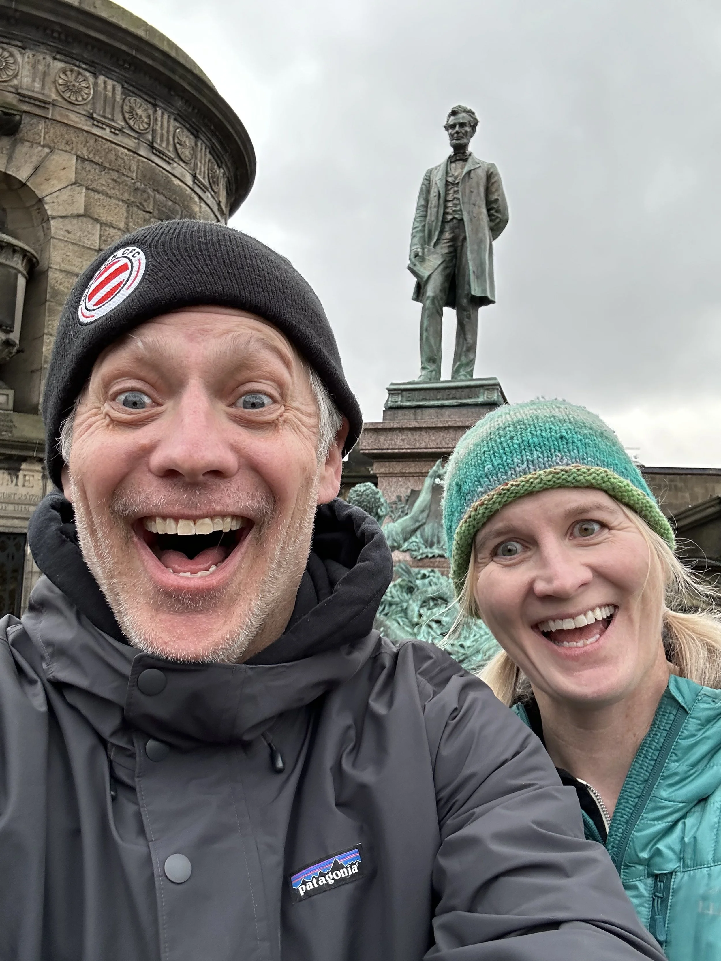 A smiling man and woman taking a selfie in front of a large statue of a man standing on a pedestal. The man is wearing a gray Patagonia jacket and a black beanie, and the woman is wearing a teal jacket and a green knit hat. The statue depicts a man d