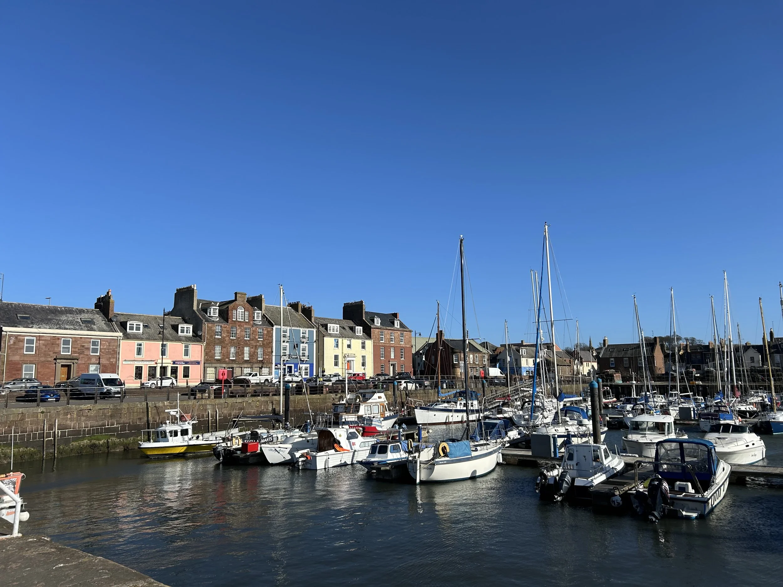 A harbor with sailboats docked in calm water, a row of colorful buildings along the waterfront, and a clear blue sky overhead.