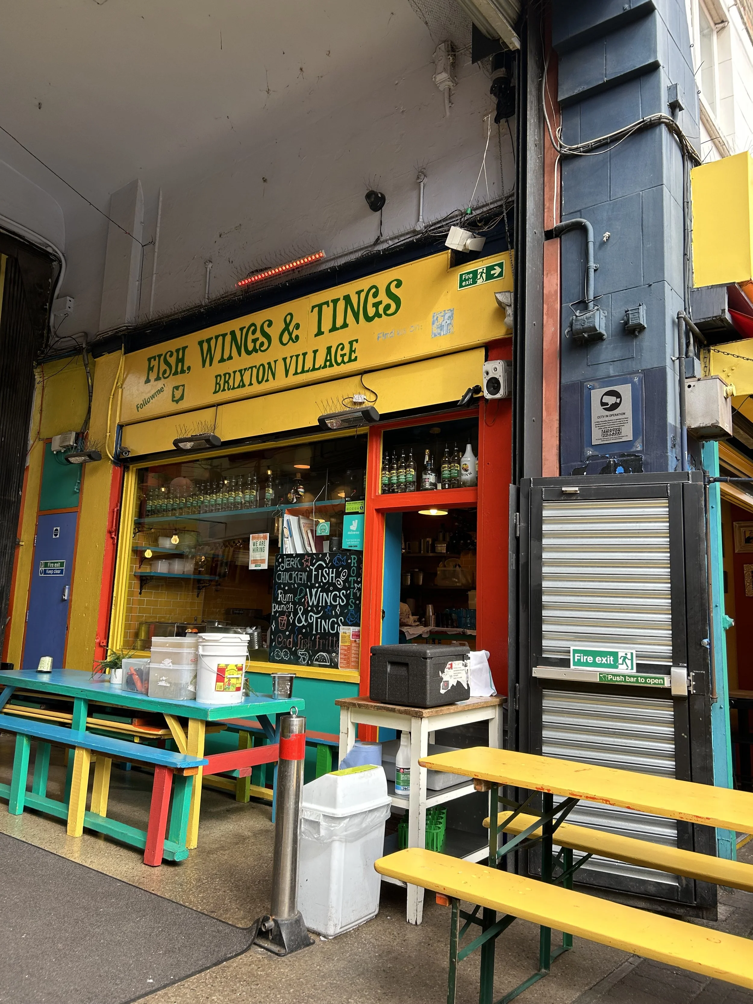 Exterior of a colorful food stand called 'Fish, Wings & Tings' in Brixton Village with outdoor seating including yellow and green benches, a chalkboard menu, and bottles displayed above the window.