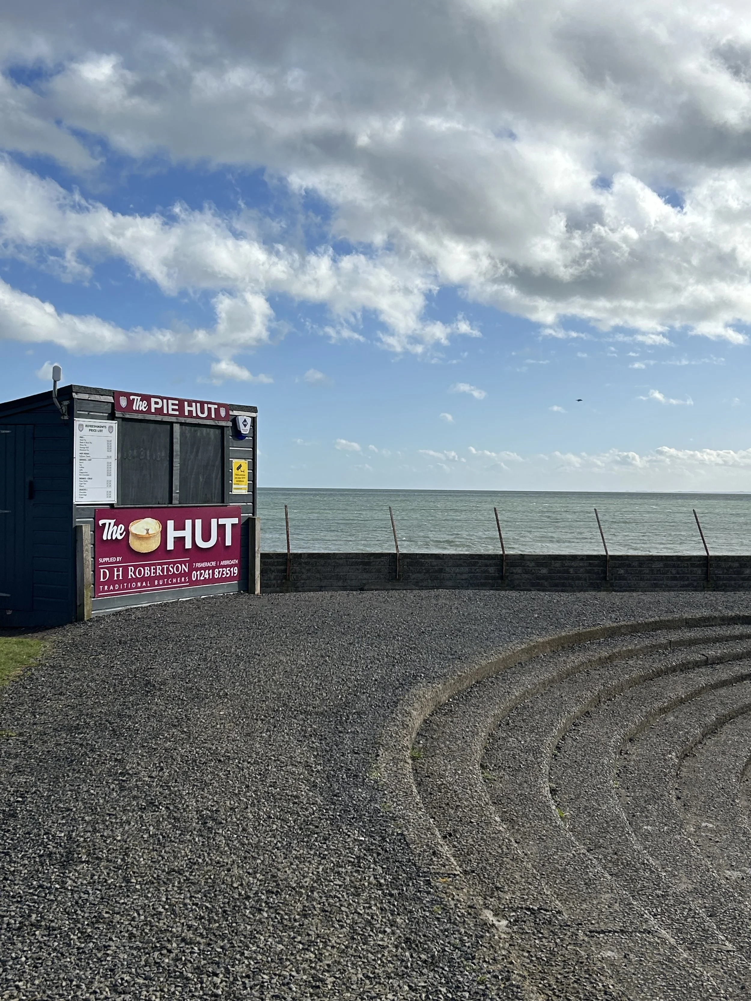 An outdoor scene by the sea showing a small blue booth with a sign that reads 'The Pie Hut,' a menu, and a phone number. The sky is partly cloudy, and there are curved park benches in the foreground along with a safety fence separating the area from 