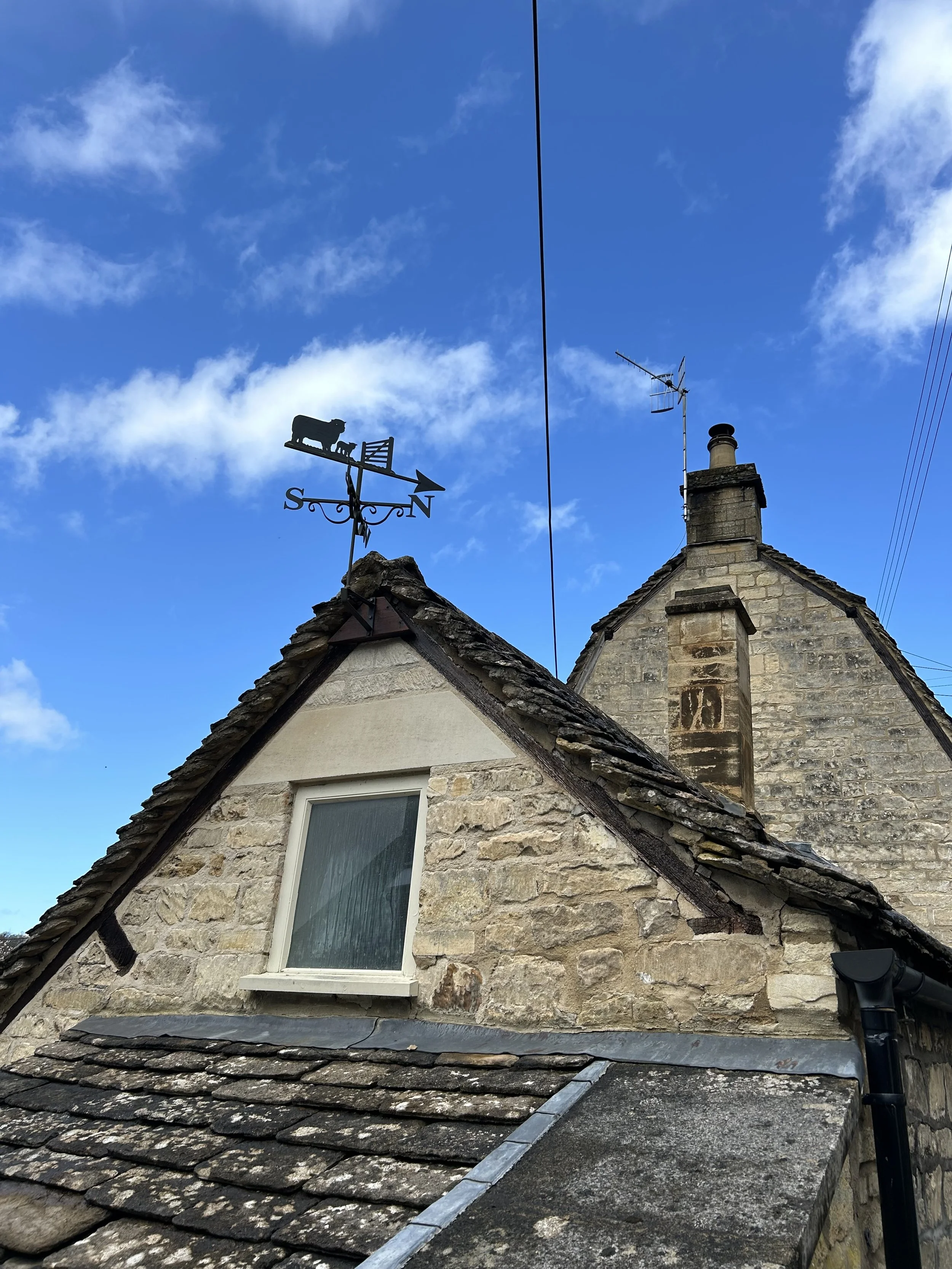 A stone house with a gabled roof and a chimney, topped with a weather vane featuring a pig, with a blue sky and clouds in the background.