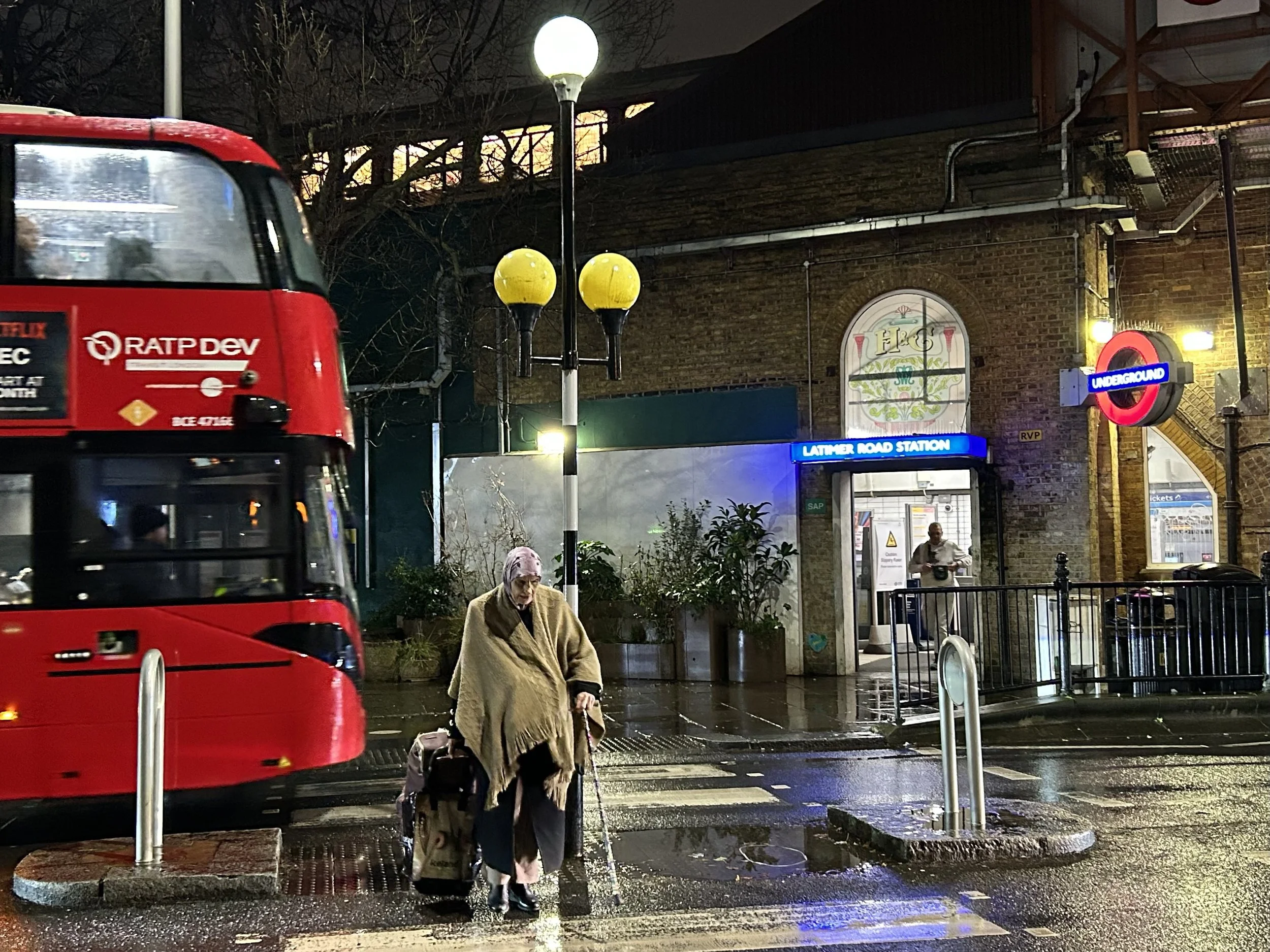 Nighttime scene of a woman with a shopping cart crossing a wet street in front of Latimer Road Station in London. A red double-decker bus passes by on the left, and a London Underground sign is visible near the station entrance. The area is illuminat