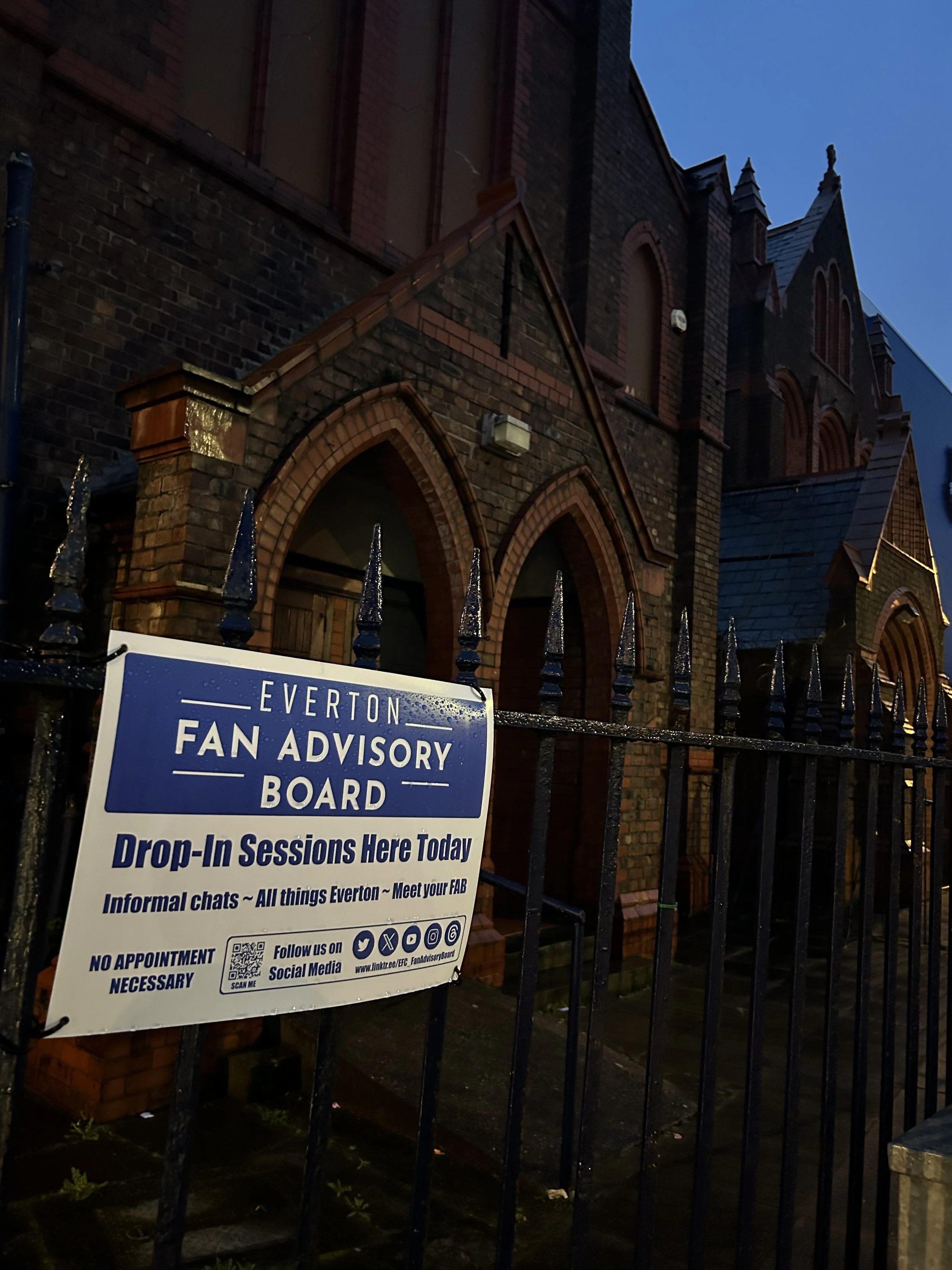 A sign on a black metal fence advertising Everton fan advisory board drop-in sessions, with a historic brick church or building in the background at dusk.