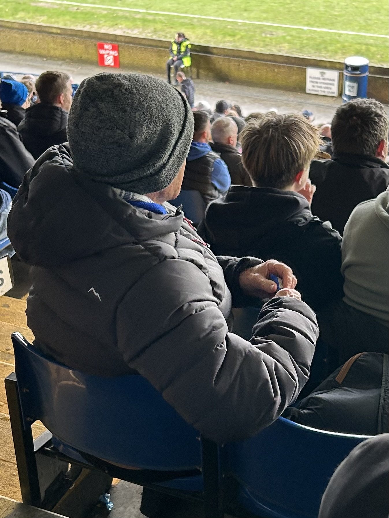 Crowd of spectators seated outdoors watching an event, with a man in a gray beanie and jacket in the foreground.