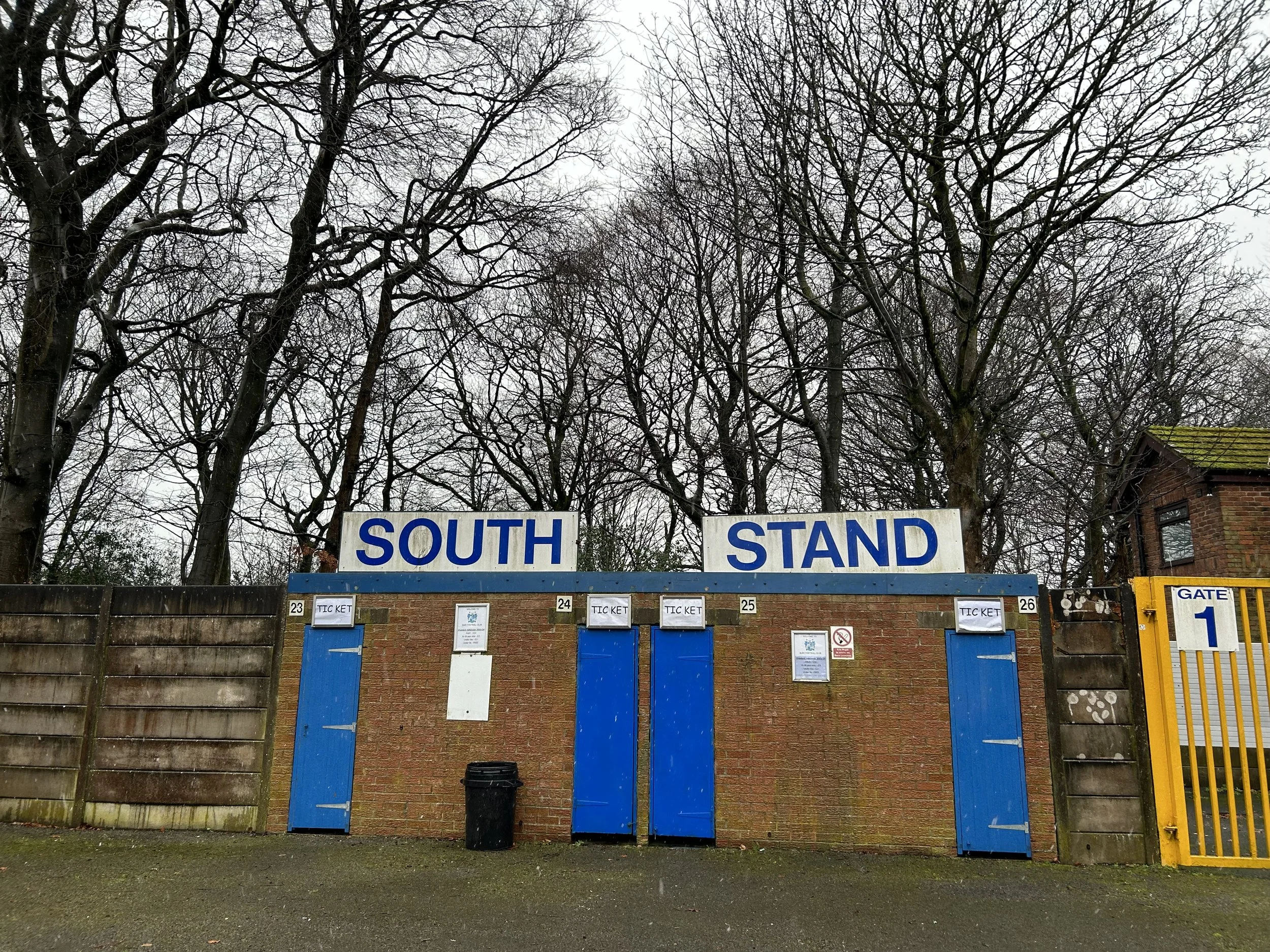 Entrance to a sports stadium with a brick wall, blue doors, and a sign reading 'SOUTH STAND', with leafless trees in the background.