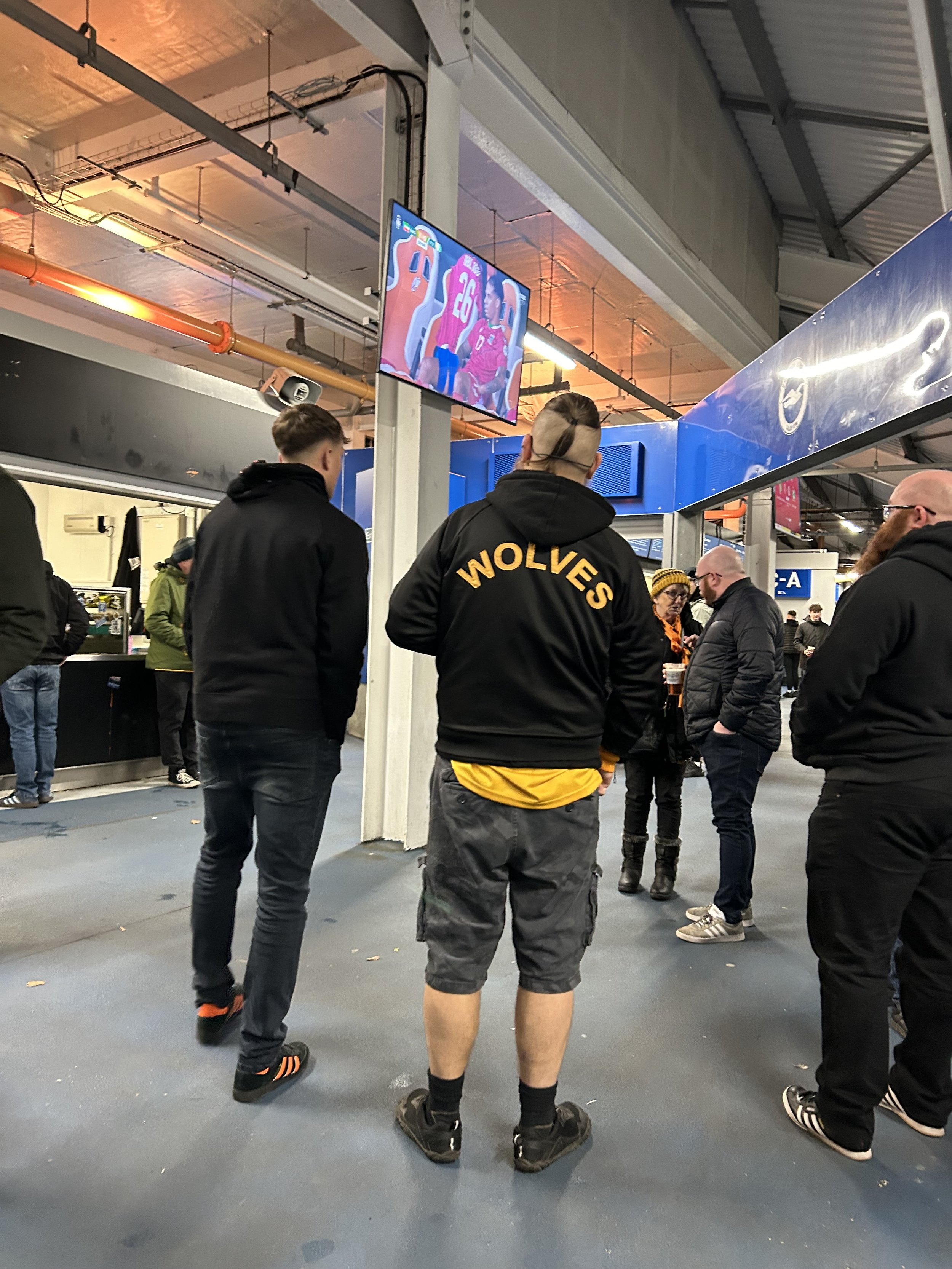 People standing in line inside an indoor arena, watching a television screen showing a hockey game, with some individuals wearing Wolverines team jackets.