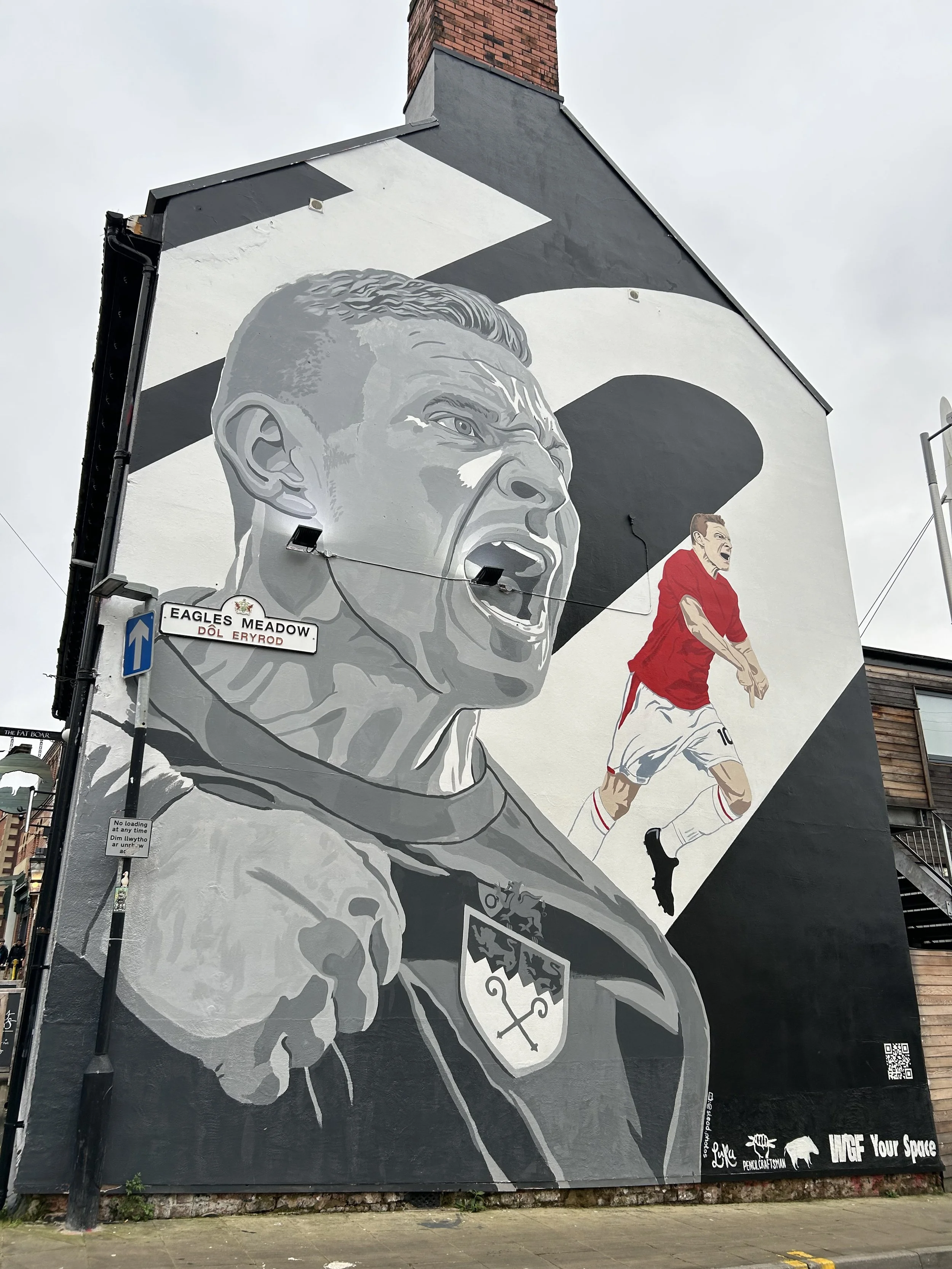 Large black and white mural of a man with a fierce expression, wearing a sports uniform with a crest, on a building wall. To the right of the mural, there is a smaller colored depiction of a soccer player in a red jersey and white shorts.